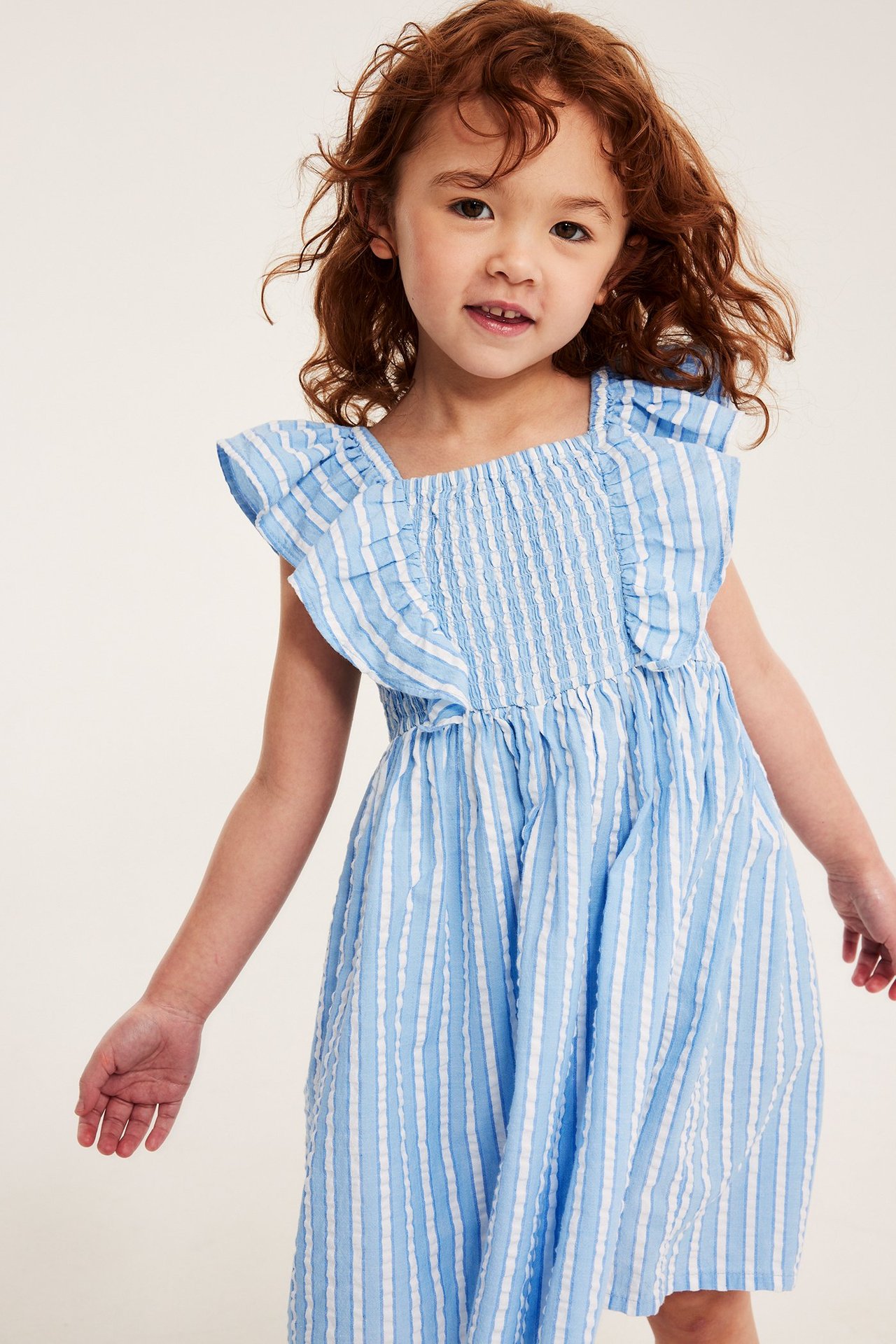 Widok z przodu, zbliżenie na dziecko w jasnoniebiesko-białej sukience z seersuckeru w paski z falbanami.
