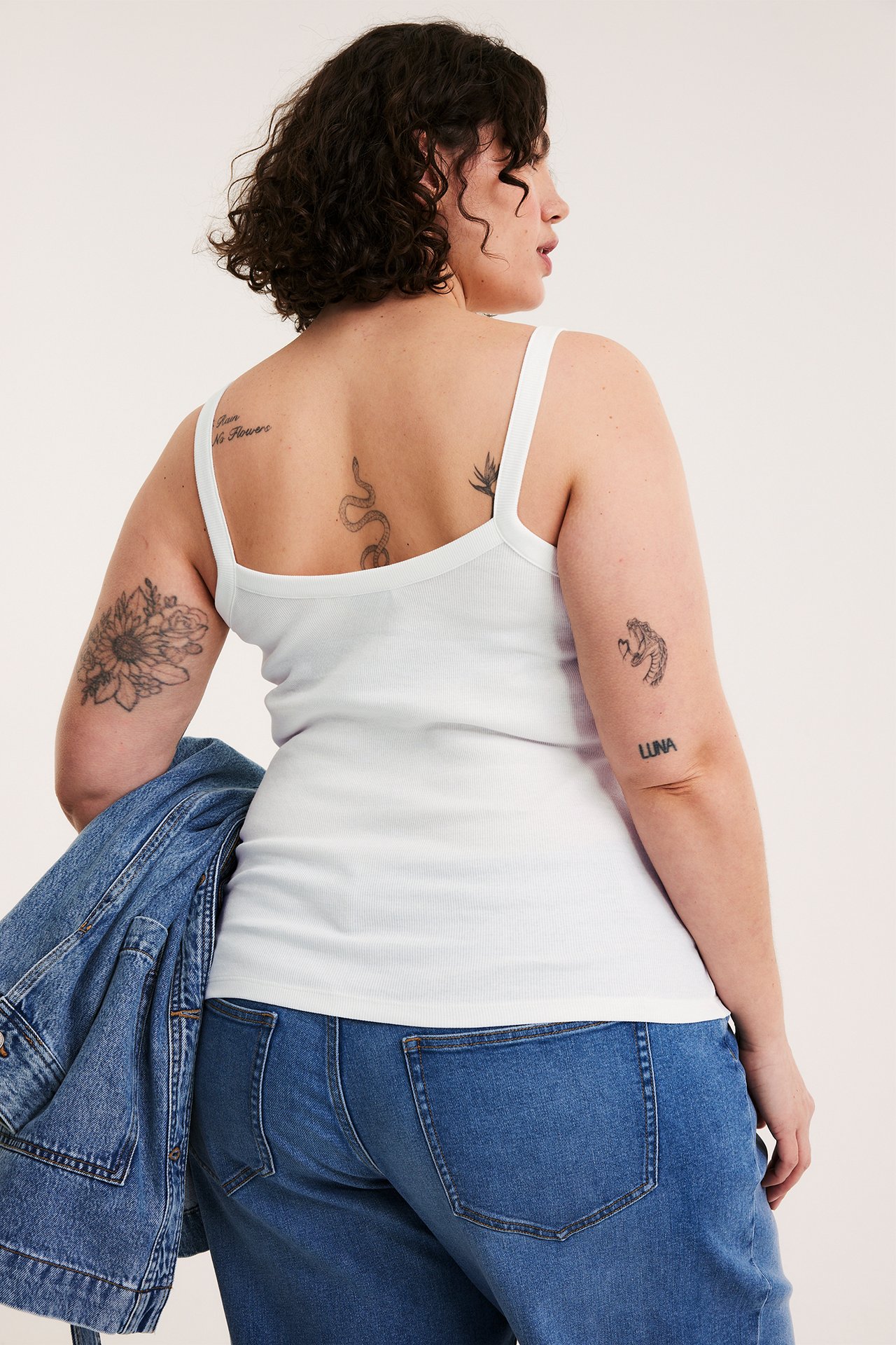 Bakfra: kvinne i hvit ribbet singlet med smale stropper, blå jeans, bærende en blå dongerijakke.