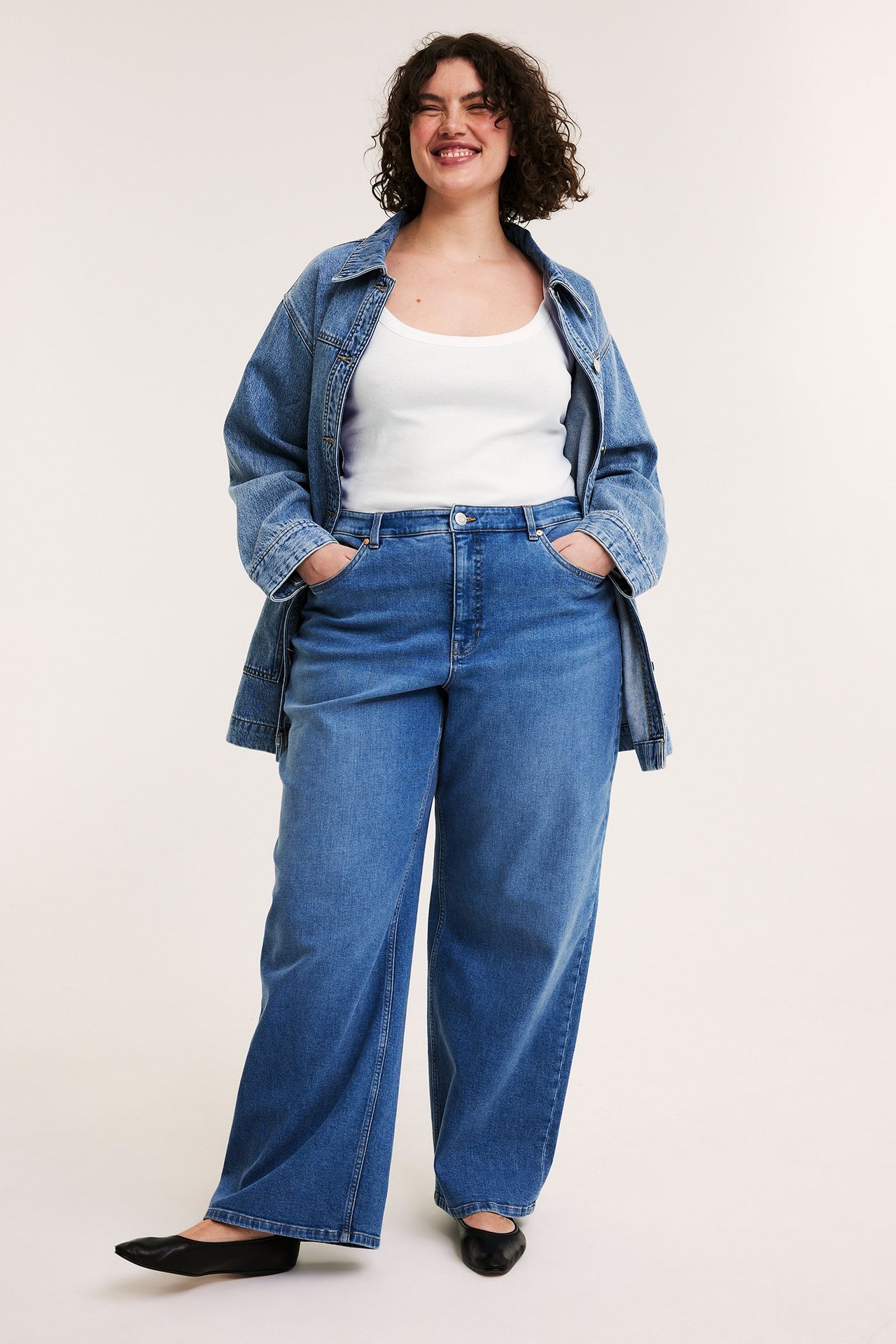 Frontbilde av kvinne i hvit ribbet singlet, åpen blå jeansjakke, vide jeans og sorte ballerinasko.