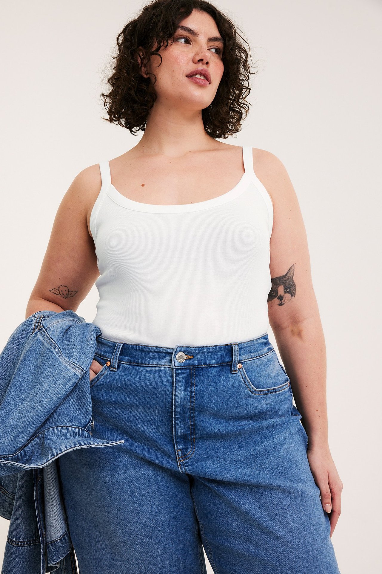 Framifrån, kvinna i vitt ribbat linne, blå jeans och blå jeansjacka hållen över armen.