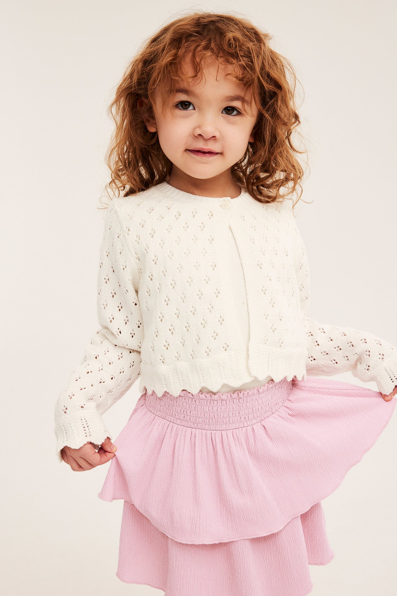 Frontvy av ett barn i ljusrosa krinklad volangkjol och naturvit stickad kofta.