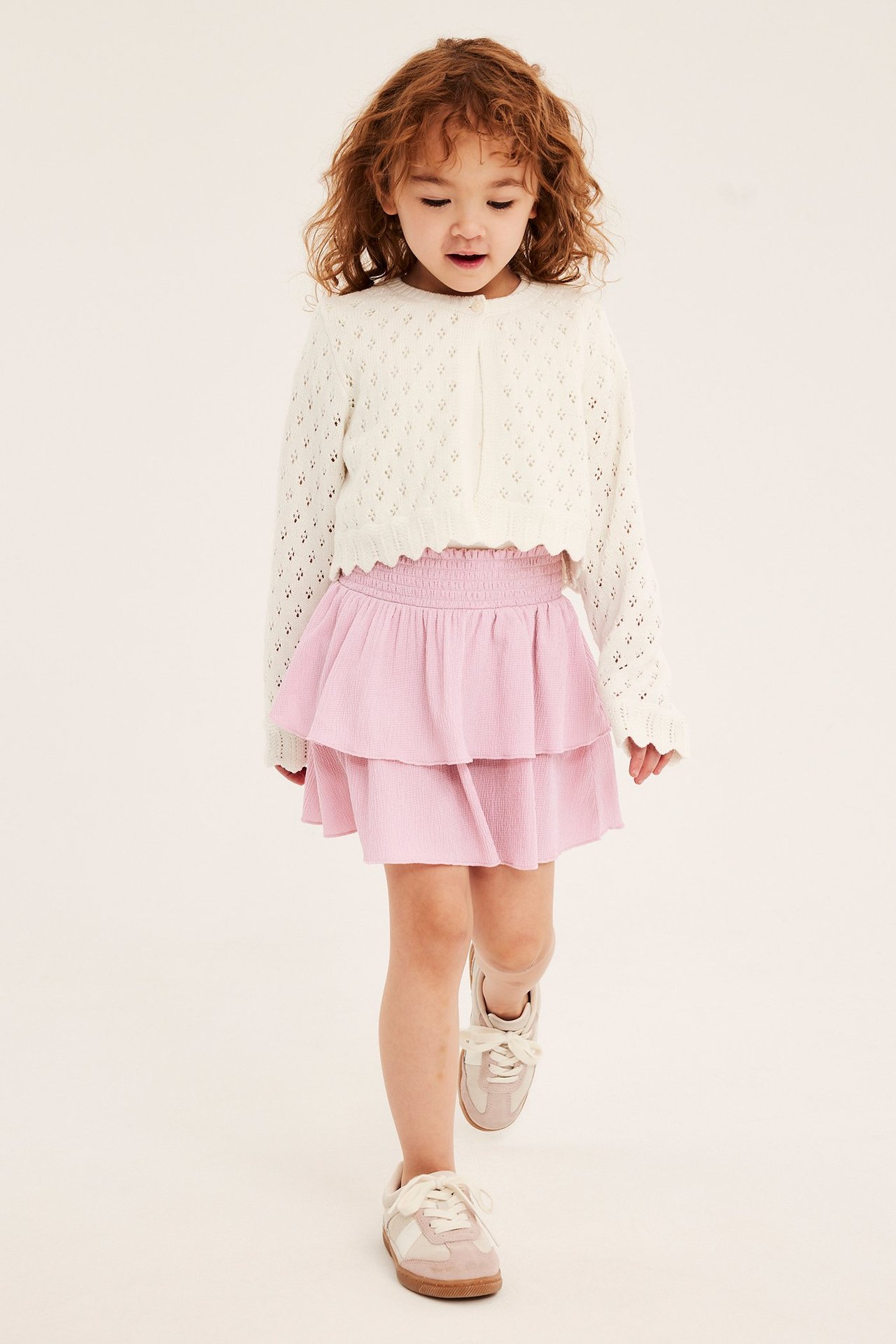 Widok z przodu: dziecko w jasnoróżowej, marszczonej spódnicy z falbanami, białym swetrze i dwukolorowych trampkach.