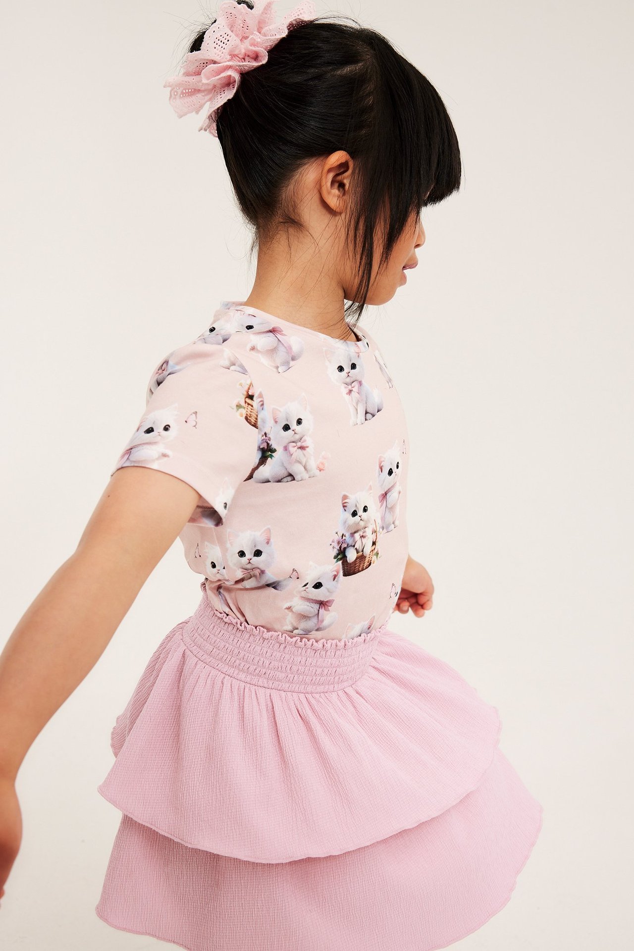 Sidovy av ett barn i ljusrosa krinklad volangkjol, en t-shirt med kattryck och en rosa scrunchie.