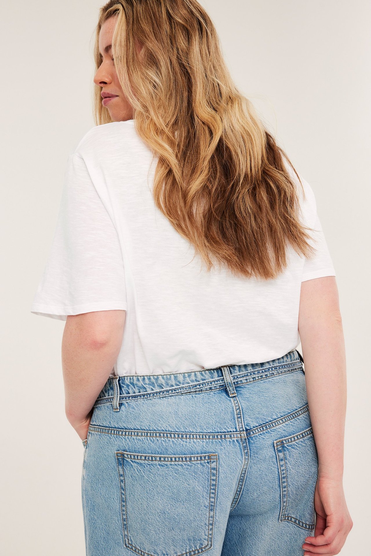 Nainen takaapäin valkoisessa V-kaula-aukko t-paidassa ja vaaleansinisissä farkuissa, käsi taskussa.