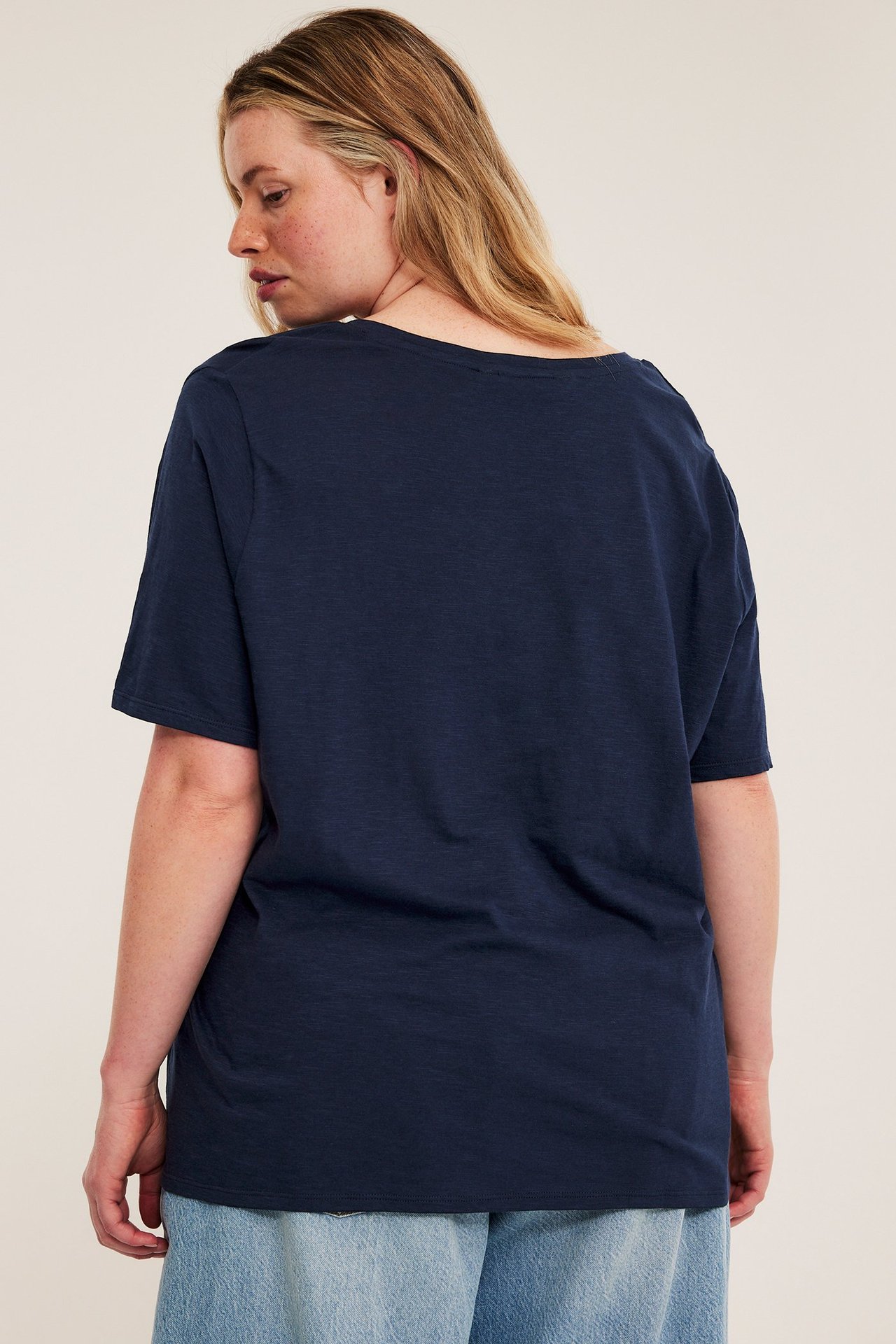 Naisen takakuva, yllään tummansininen, rento puuvillainen V-aukkoinen t-paita ja vaaleansiniset farkut.