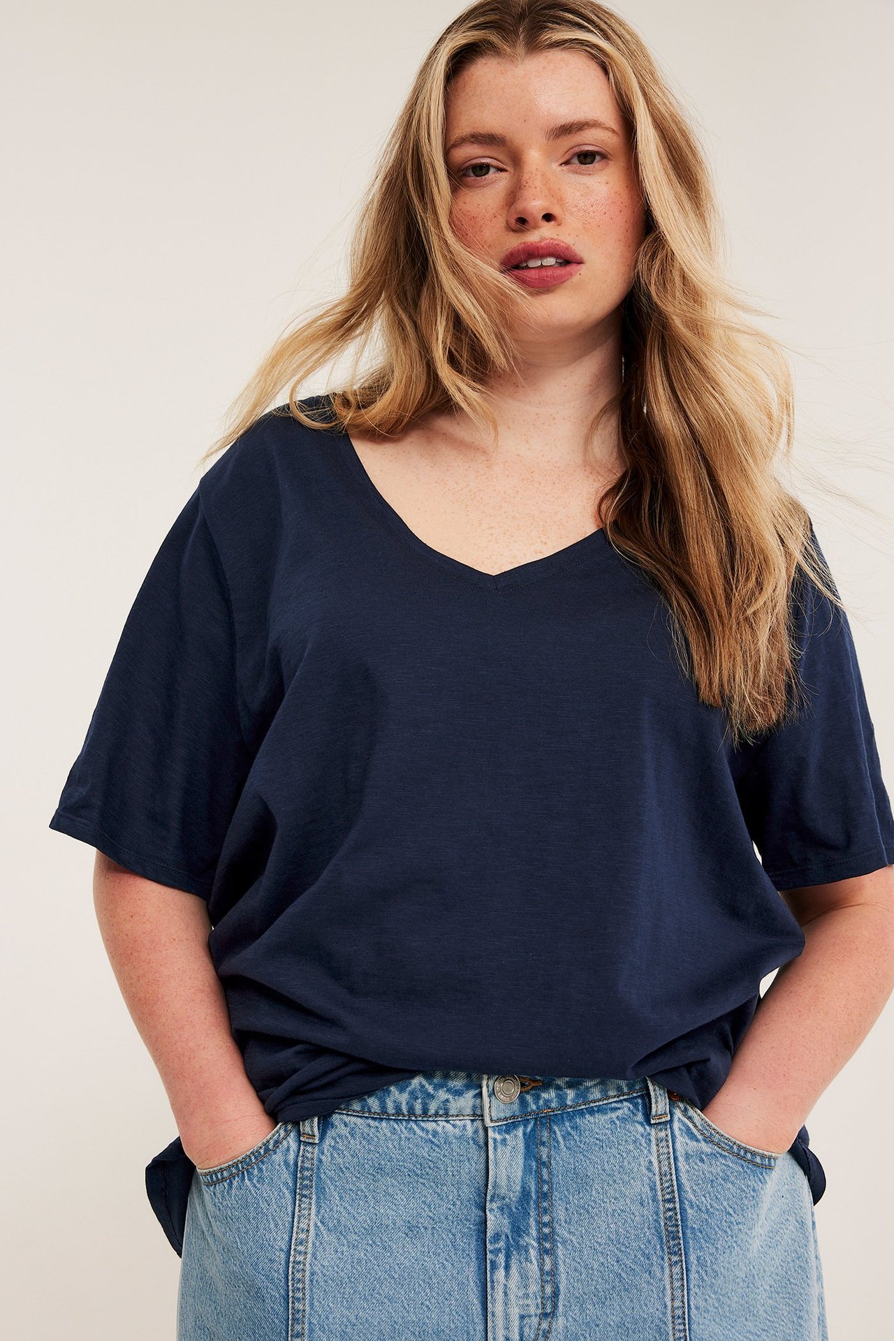 Edestä, nainen tummansinisessä V-kaula-aukkoisessa puuvillaisessa T-paidassa ja vaaleissa farkuissa, kädet taskuissa.