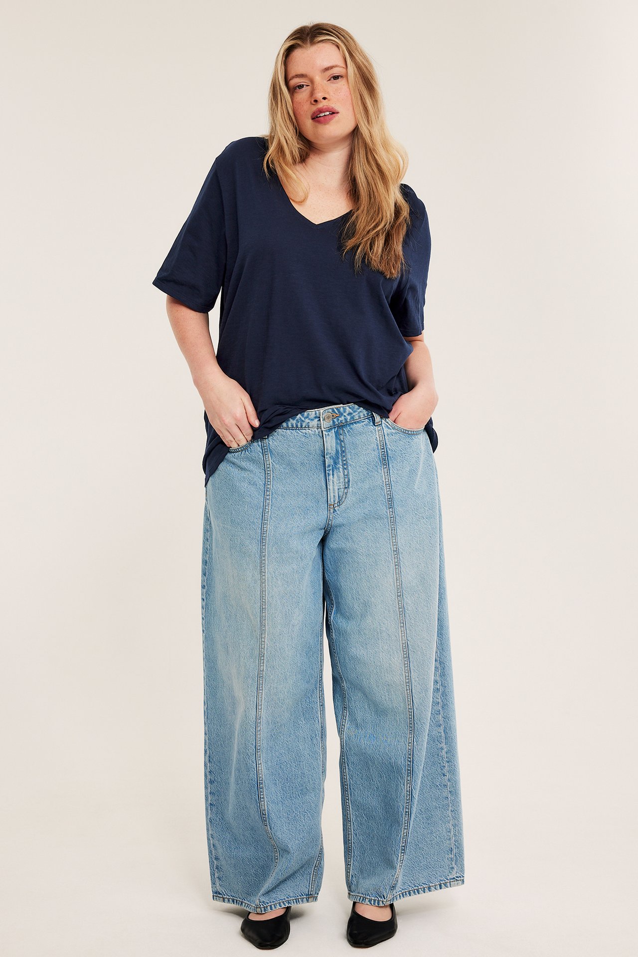 Edestä: Nainen tummansinisessä V-pääntien puuvillapaidassa, vaaleansinisissä leveissä farkuissa, mustissa kengissä.