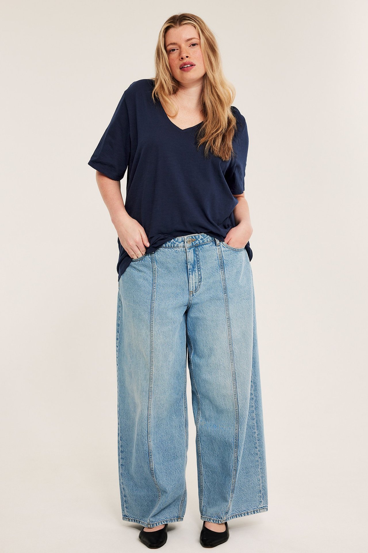 Edestä: Nainen tummansinisessä V-pääntien puuvillapaidassa, vaaleansinisissä leveissä farkuissa, mustissa kengissä.