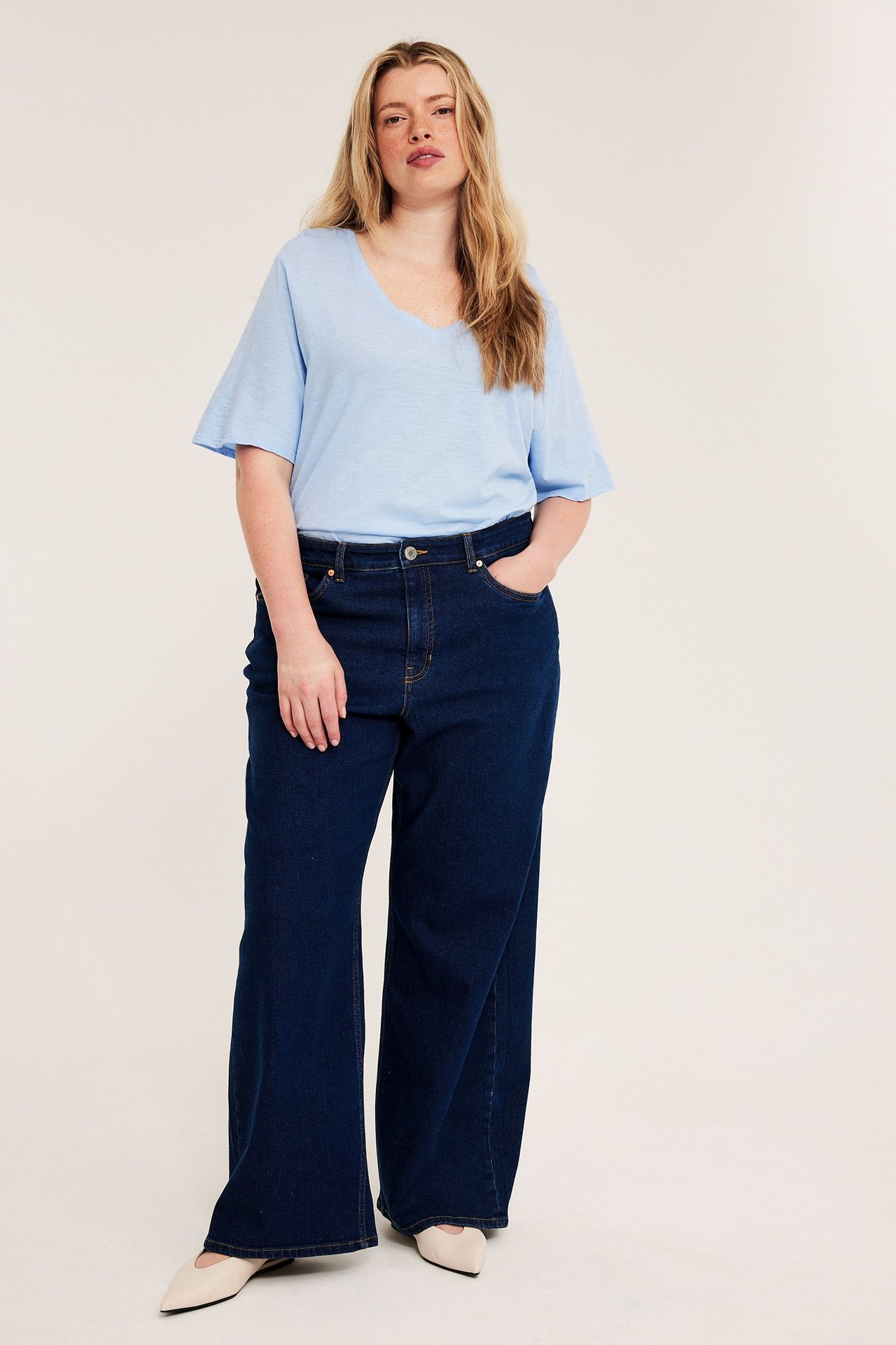 Edestä, nainen vaaleansinisessä V-aukkopuuvillapaidassa, tummansinisissä leveälahkeisissa farkuissa ja beigeissä avokkaissa.