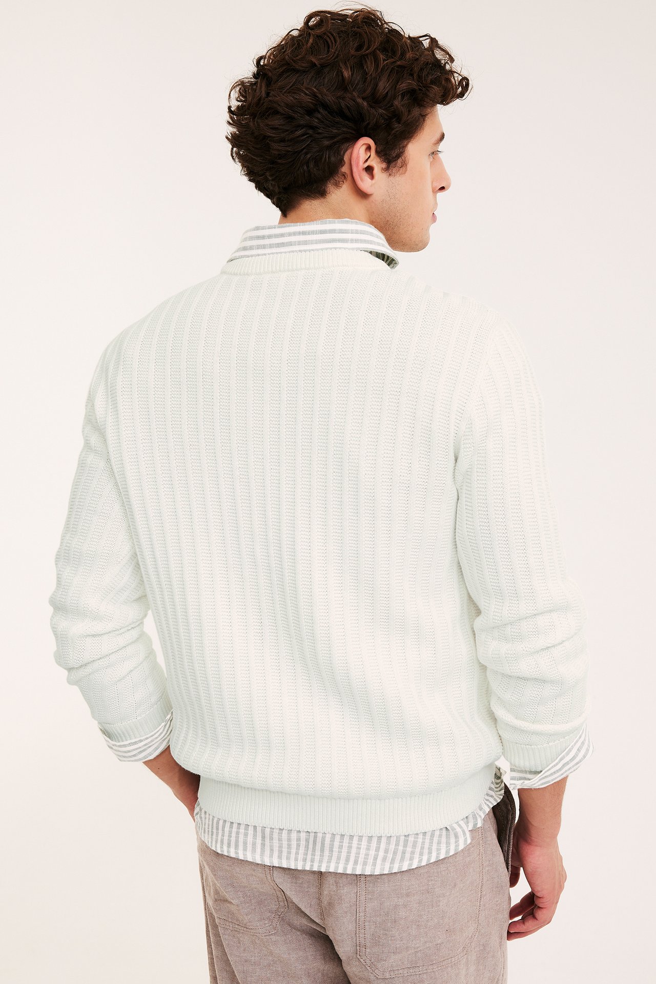 Mężczyzna, od tyłu, w kremowym swetrze o strukturalnym splocie, koszuli w paski i beżowych spodniach.