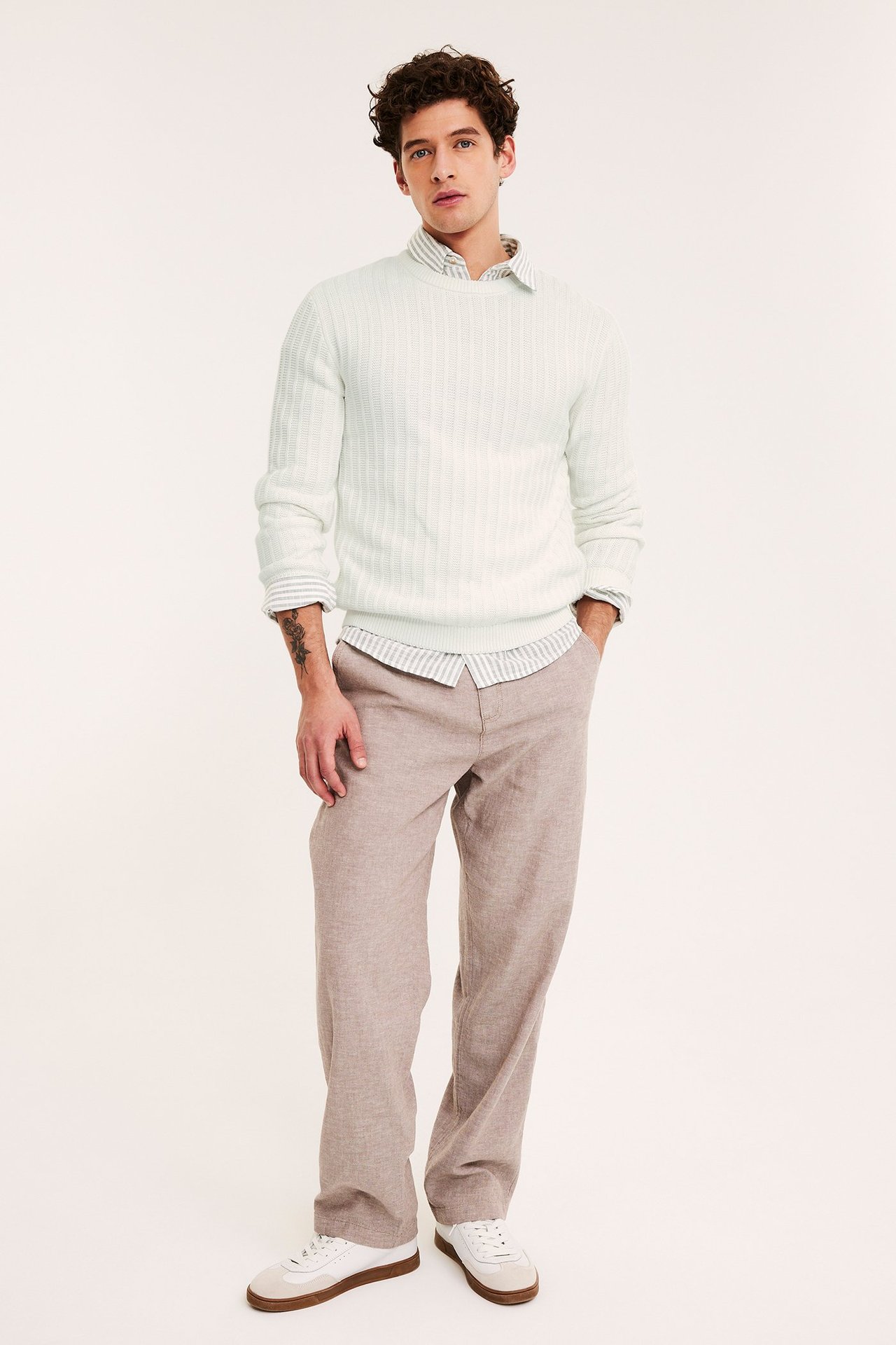 Edestäpäin mies luonnonvalkoisessa struktuurineulepuserossa, raidallisessa paidassa, vaaleanruskeissa housuissa ja valkoisissa tennareissa.