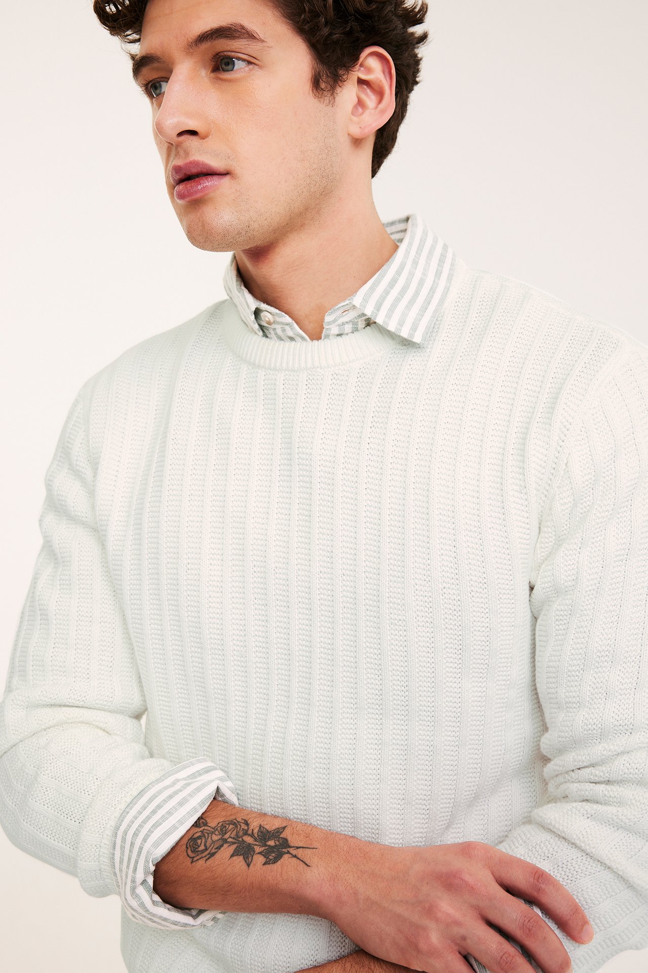 Zbliżenie mężczyzny w ecru strukturalnym swetrze z okrągłym dekoltem, na szarej koszuli w paski.