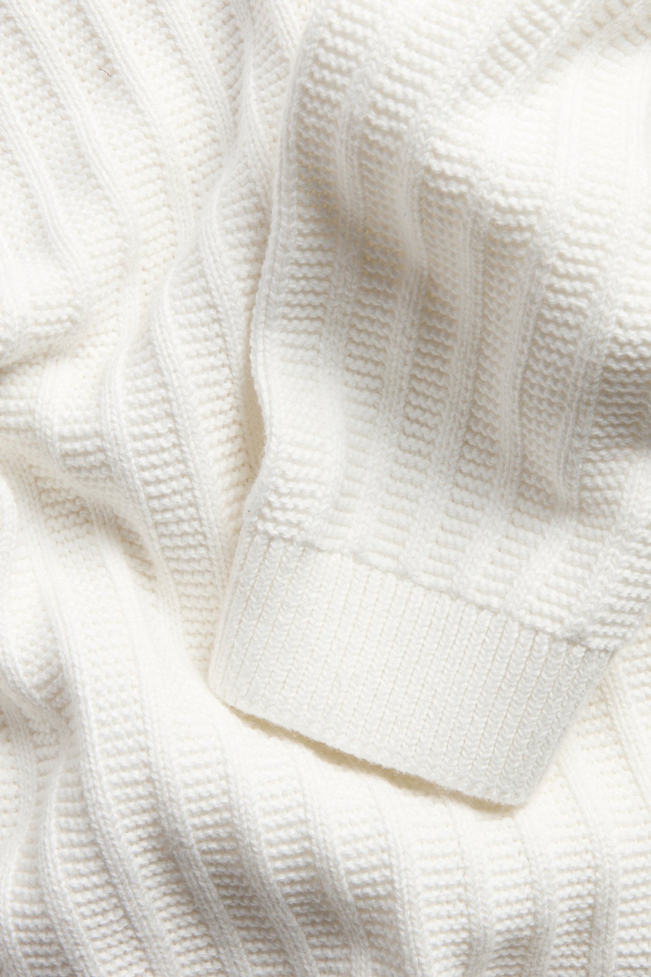Nærbilde av en offwhite bomulls strikkegenser med strukturmønster og ribbestrikket mansjett.