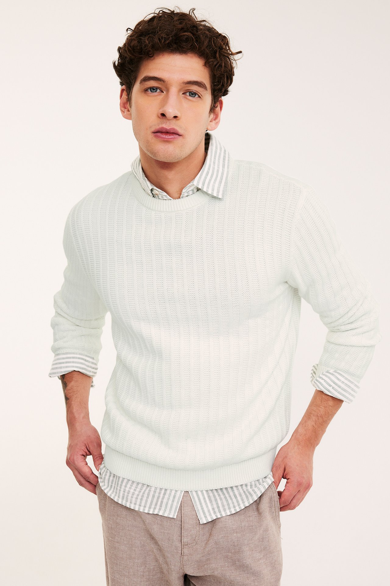 Edestä: Mies luonnonvalkoisessa struktuurineuleessa raidallisen paidan ja vaaleanruskeiden housujen kanssa.