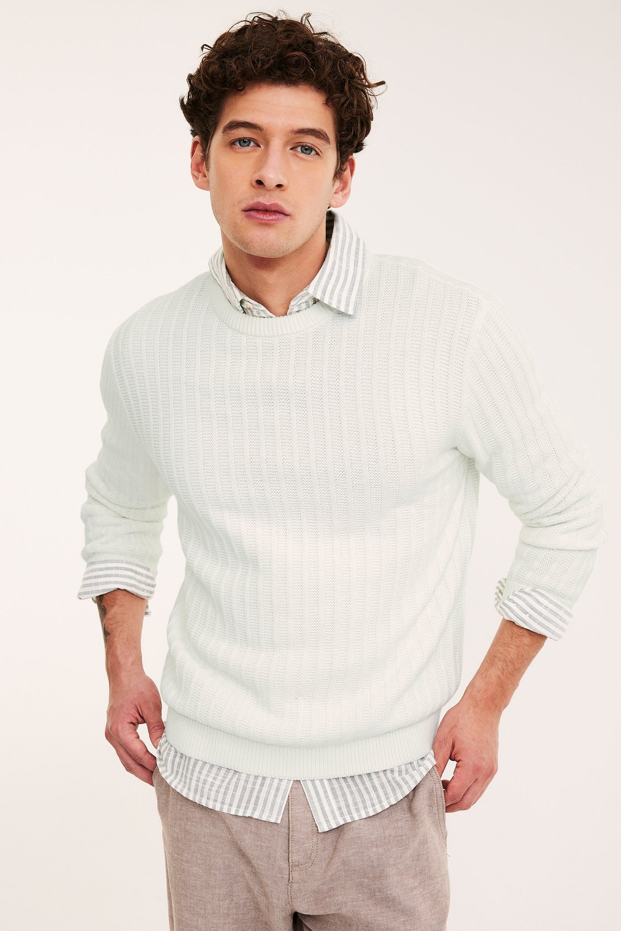 Widok z przodu: Mężczyzna w ekri swetrze o strukturze splotu, na koszuli w paski i beżowych spodniach.