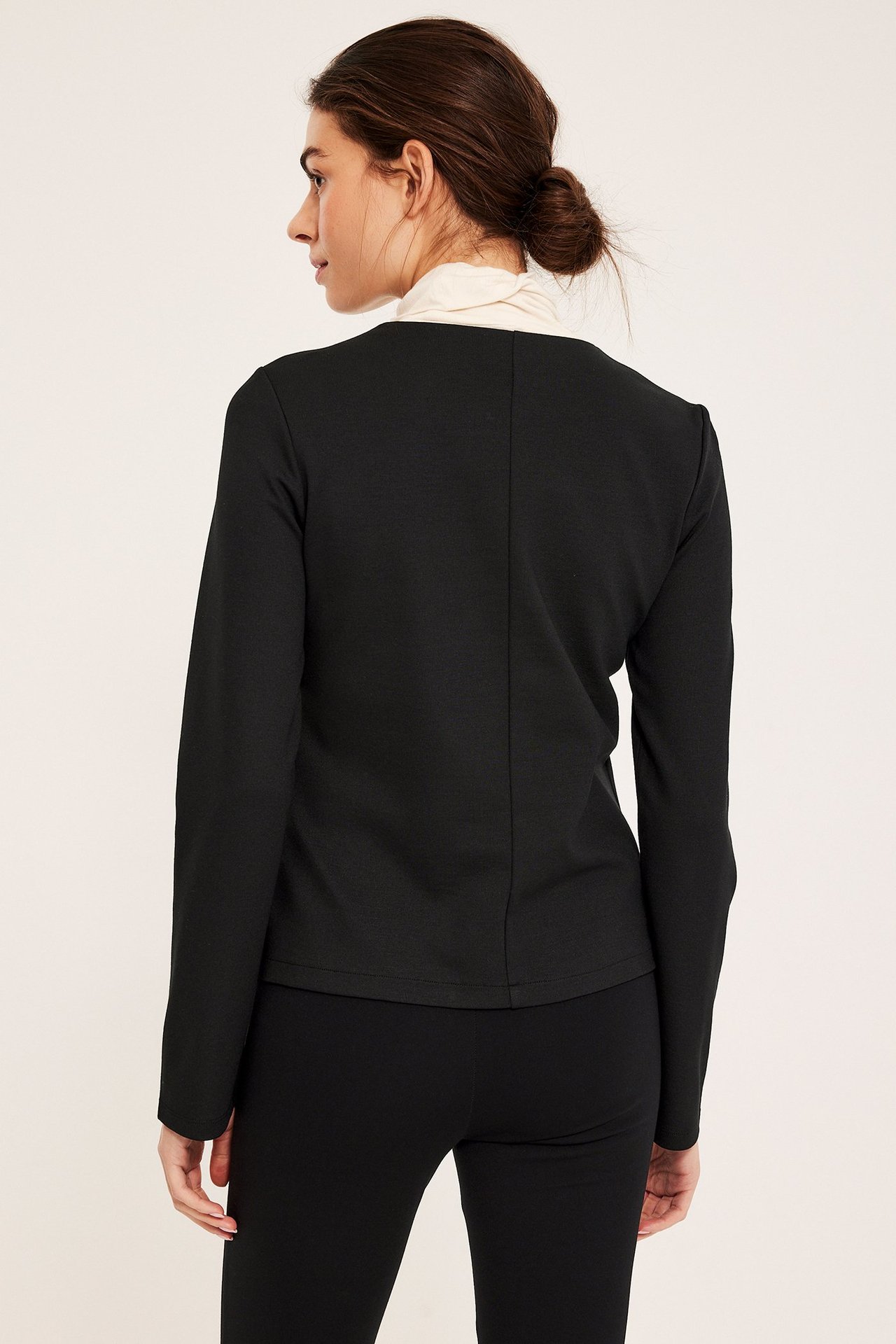 Naisen selkäkuva mustassa tyköistuvassa takissa vaalean poolopaidan päällä, yhdistettynä mustiin housuihin.