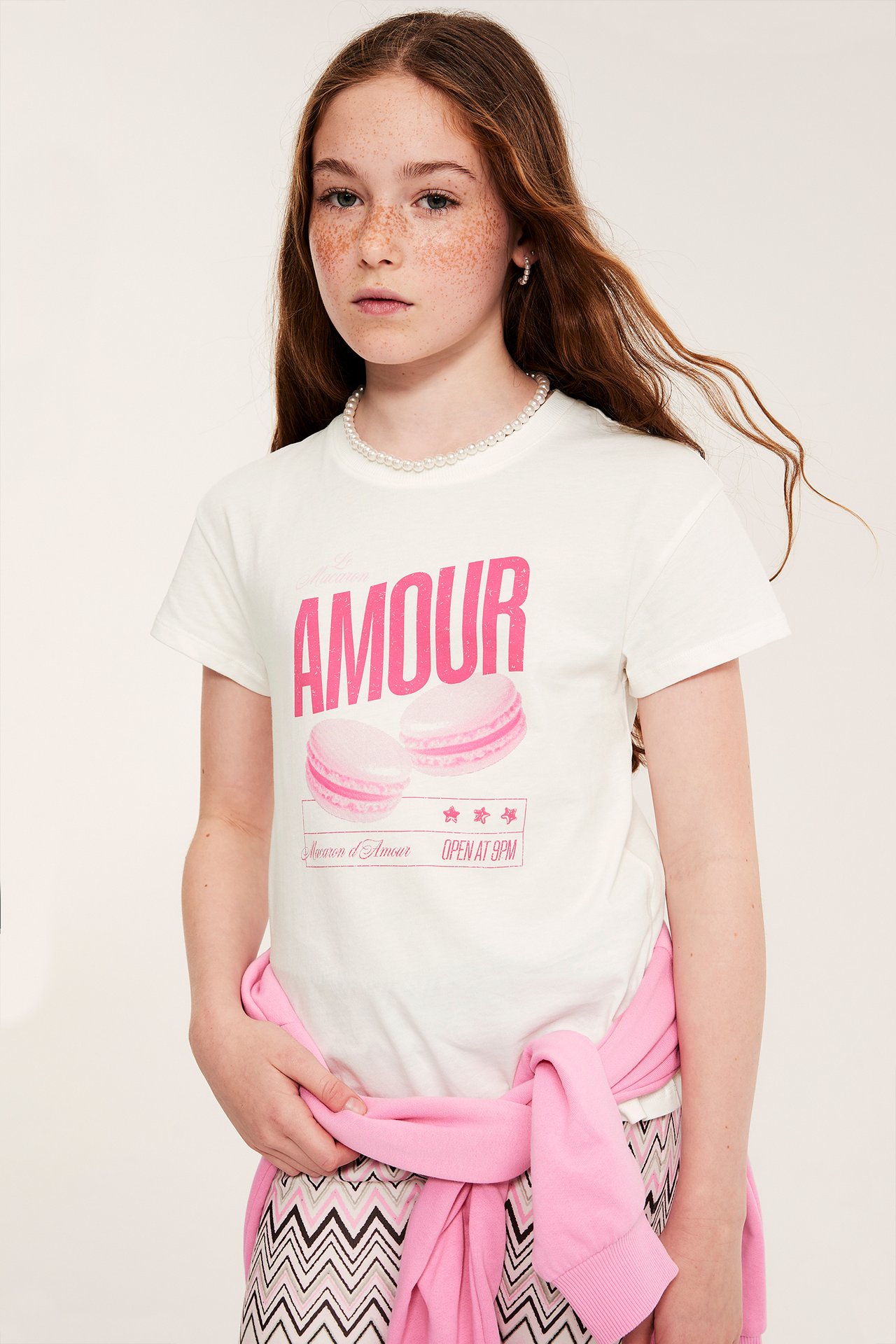 Widok z przodu: dziecko w białym bawełnianym T-shircie z różowym napisem 'AMOUR', różowy sweter w pasie, wzorzyste spodnie.
