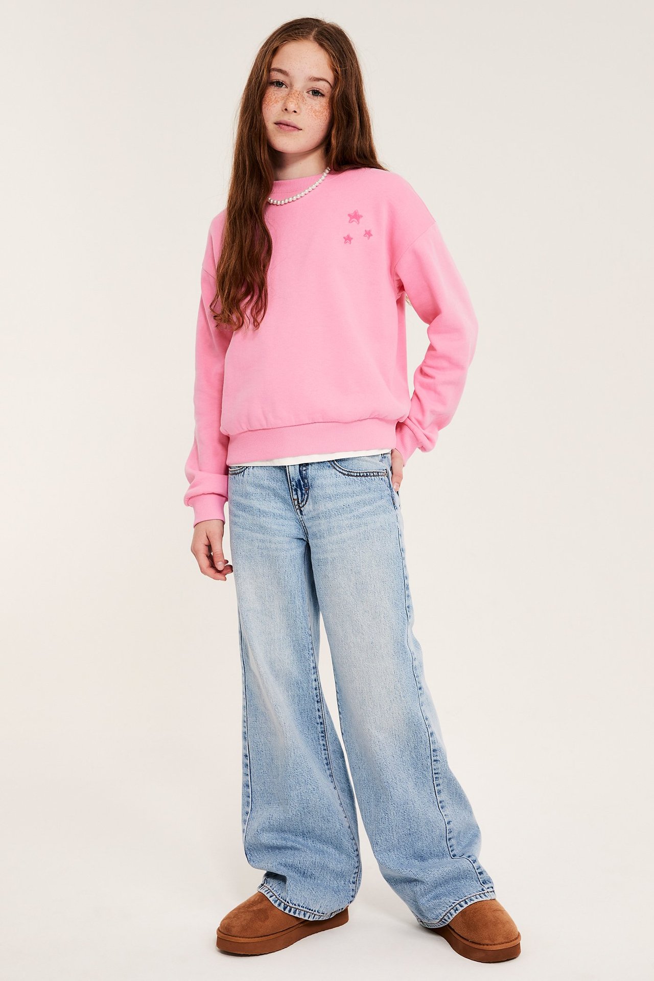 Edestä: lapsi vaaleanpunaisessa tähtipaidassa, vaaleansinisissä farkuissa, ruskeissa platform-kengissä, helmikaulakorussa.