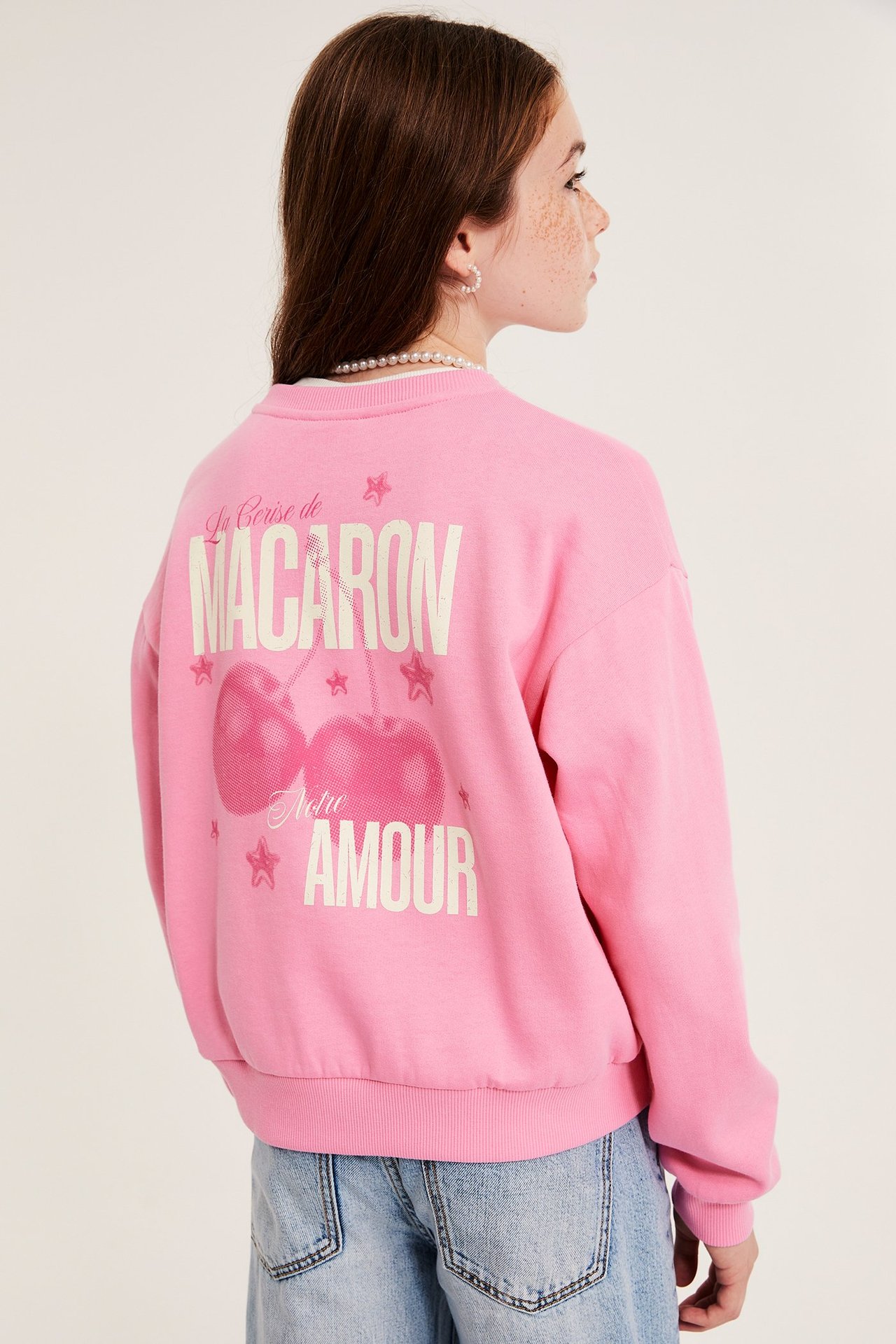 Lapsi takaa päin, yllään vaaleanpunainen kuvioitu collegepaita, vaaleansiniset farkut, helmikaulakoru, korvakorut.