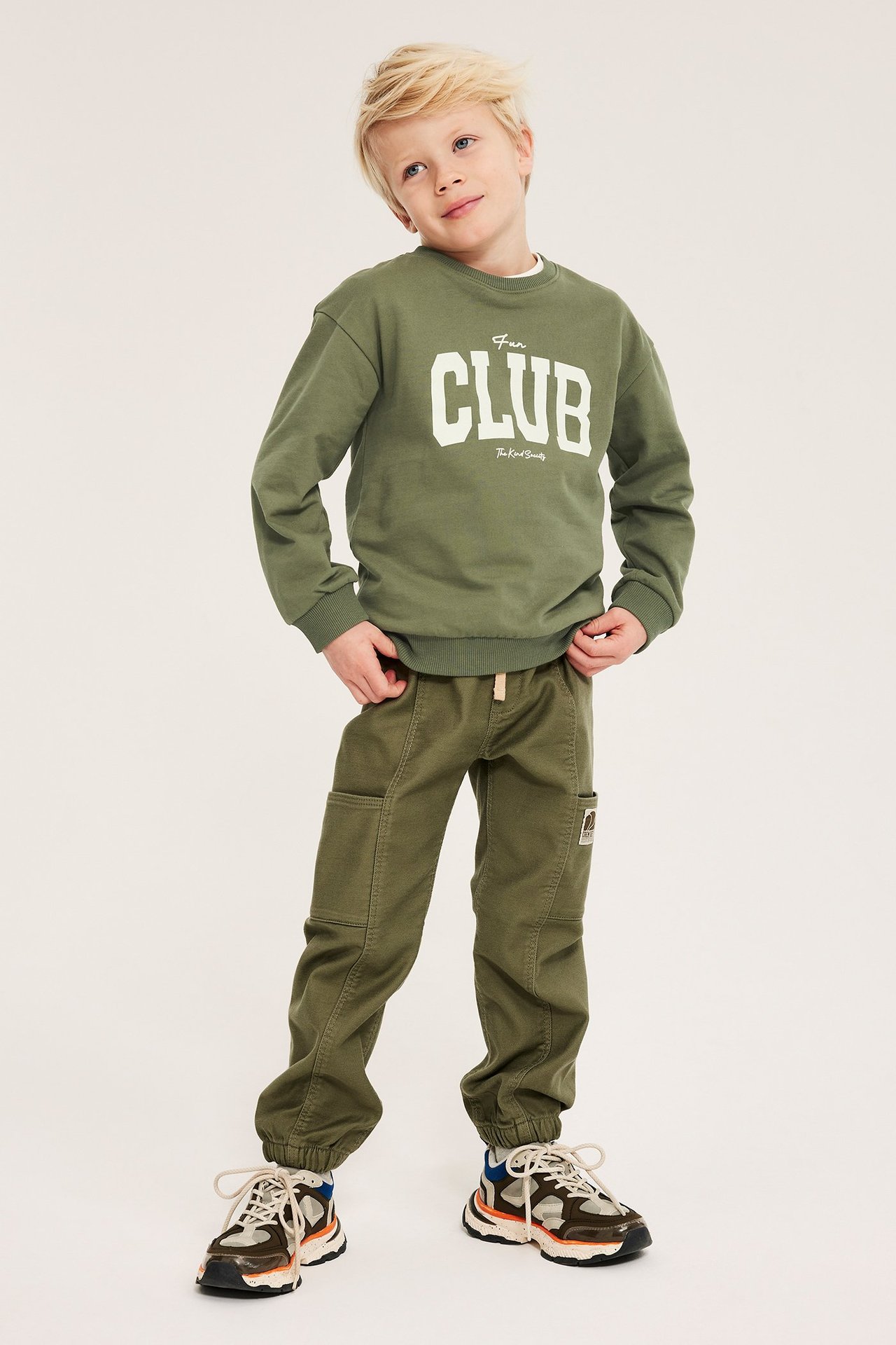 Forfra: gutt i mørkegrønn 'CLUB'-genser, grønne cargobukser og flerfargede joggesko.