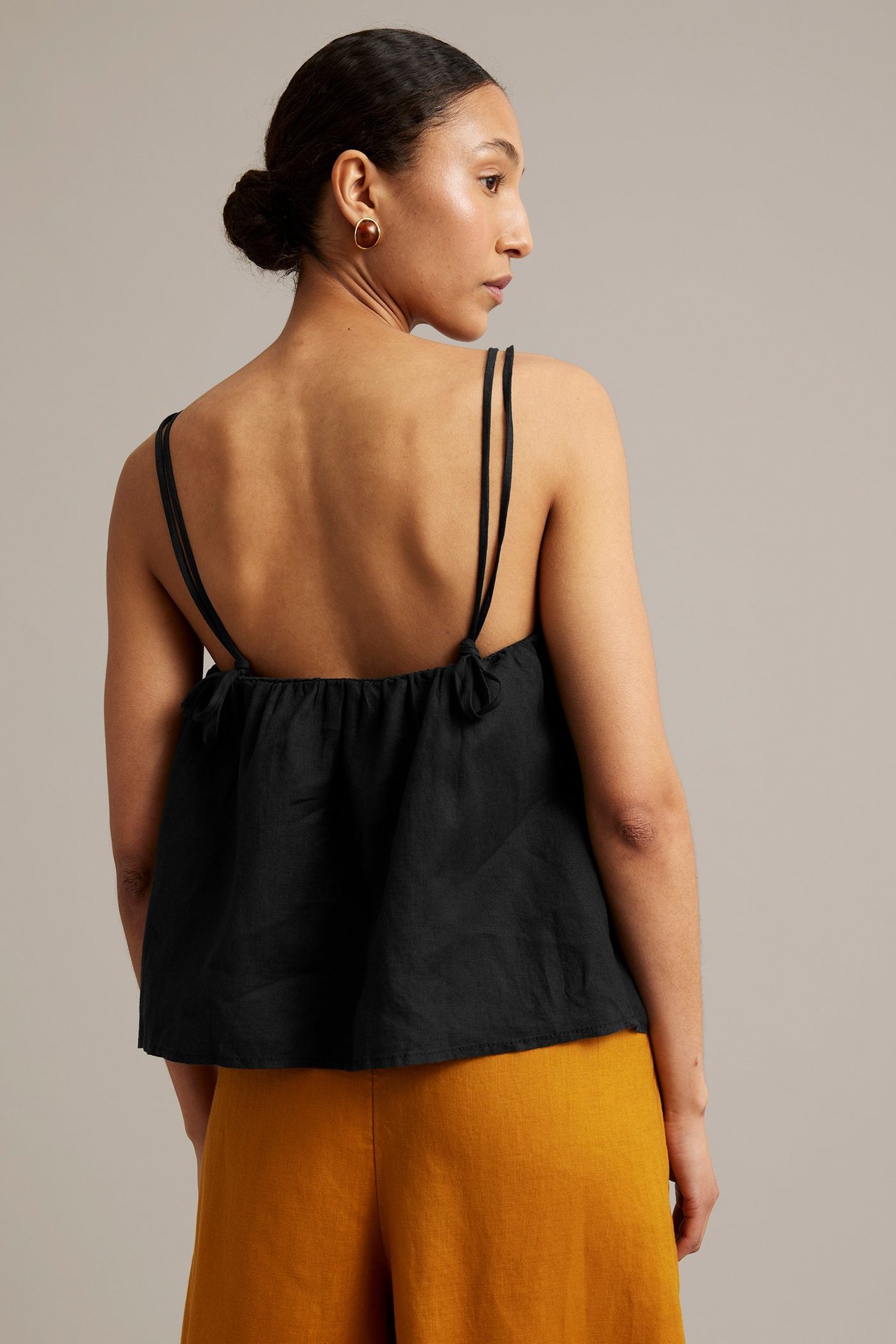 Bakifrån, kvinna i svart linne av linne med smala knytband och gula vida byxor, med bruna örhängen.