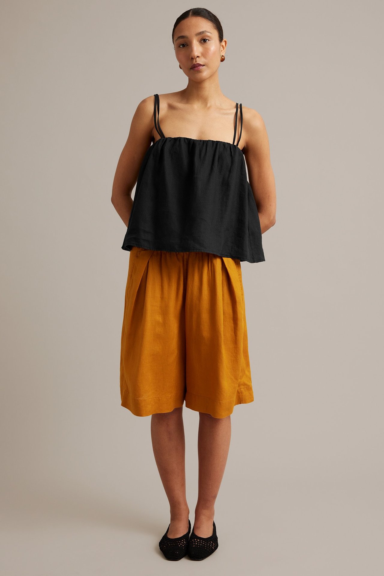 Forfra: kvinne i svart linne med smale stropper og okerfargede plisserte shorts.