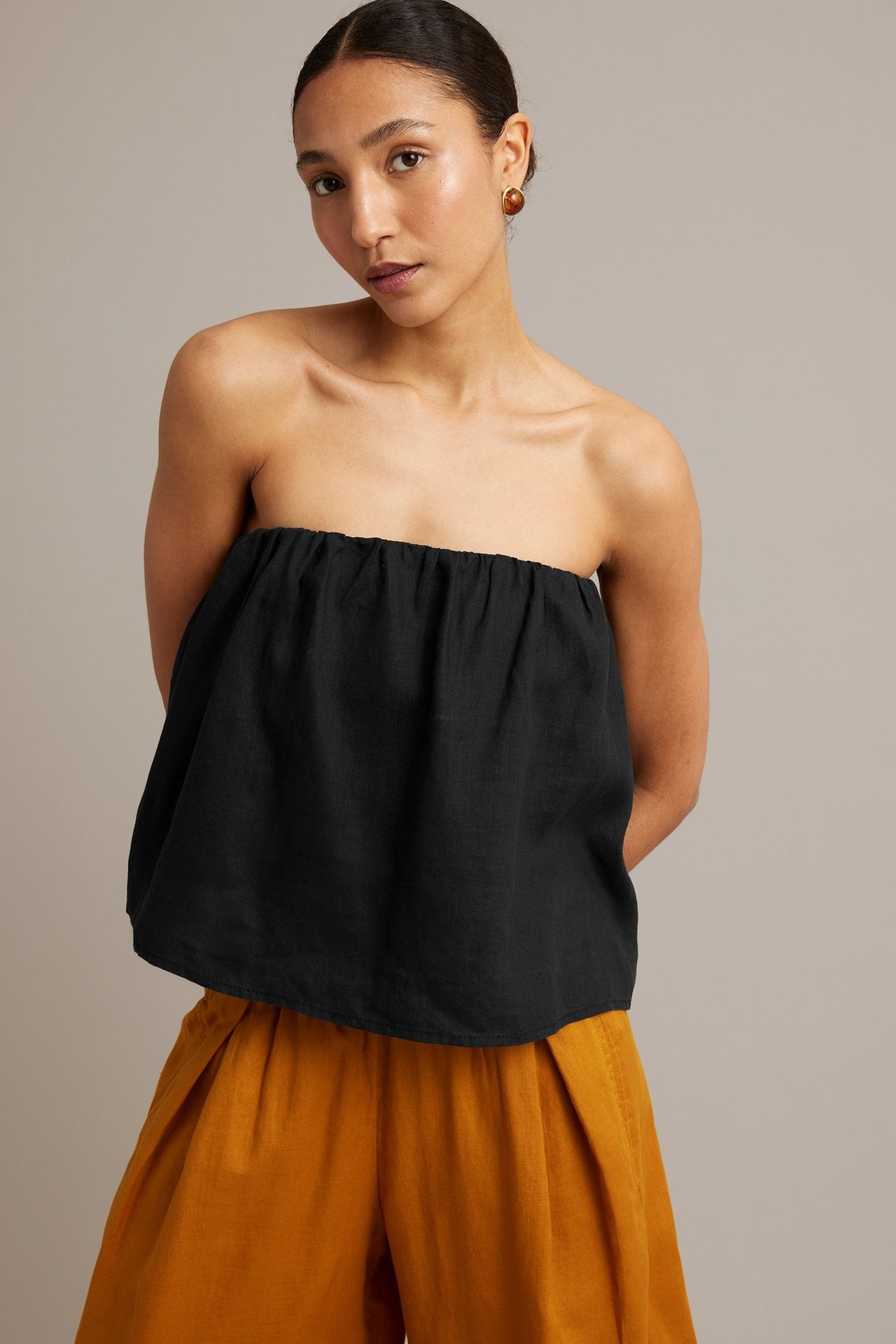 Frontbilde av en kvinne i svart stroppeløs lintopp, oransje vide bukser og ravfargede øredobber.