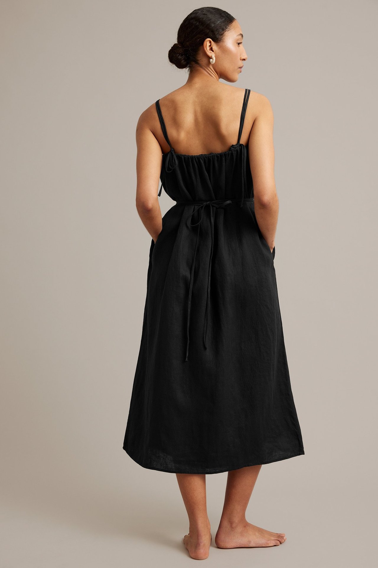Bakifrån: kvinna bär en svart linneklänning med smala axelband och knytbälte i midjan.