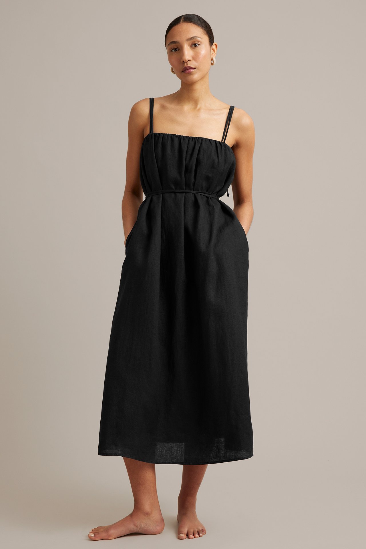 Framifrån, en kvinna i svart linneklänning med smala axelband och knytskärp.