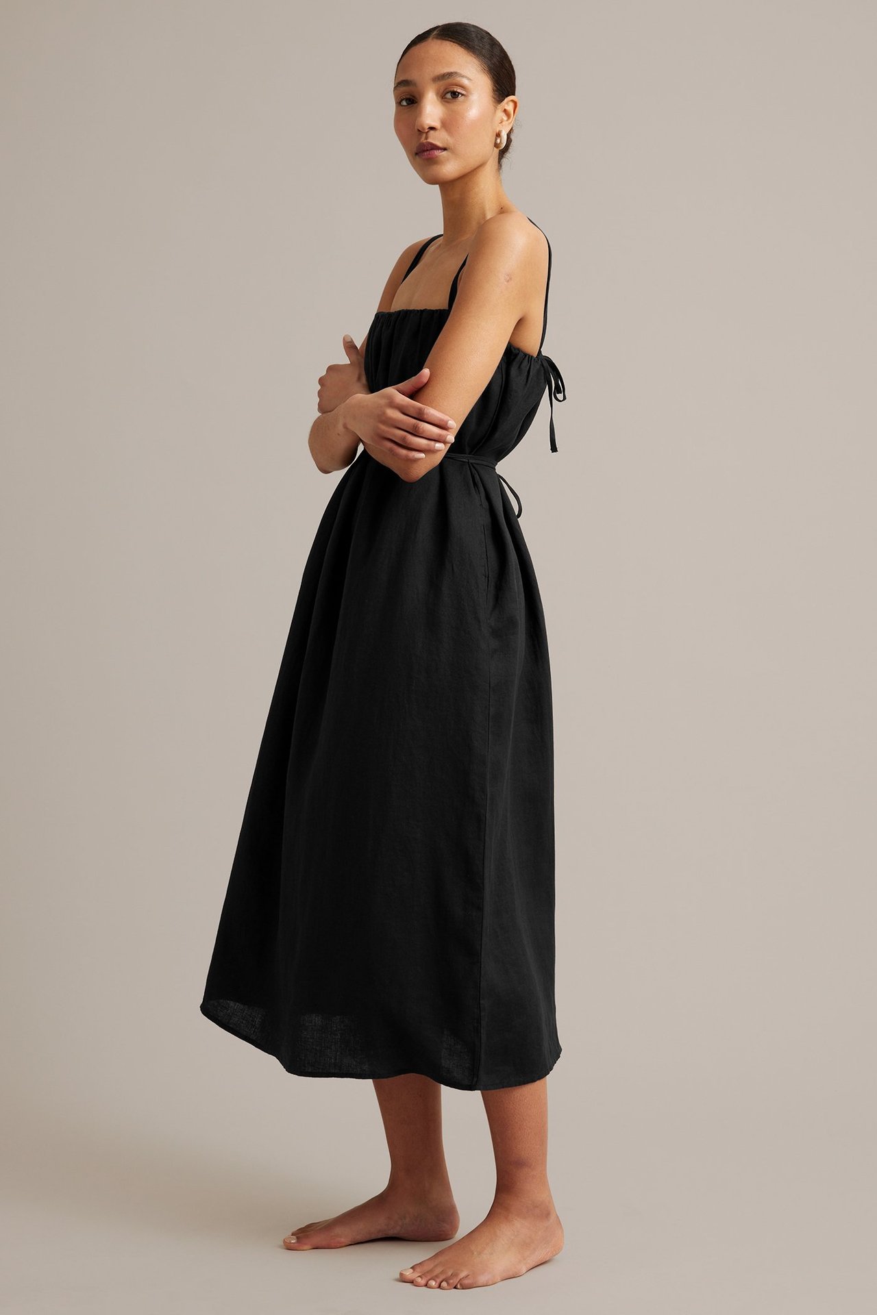 Sidovy av kvinna i svart linneklänning med knytband och midjebälte, bärande ringörhängen.