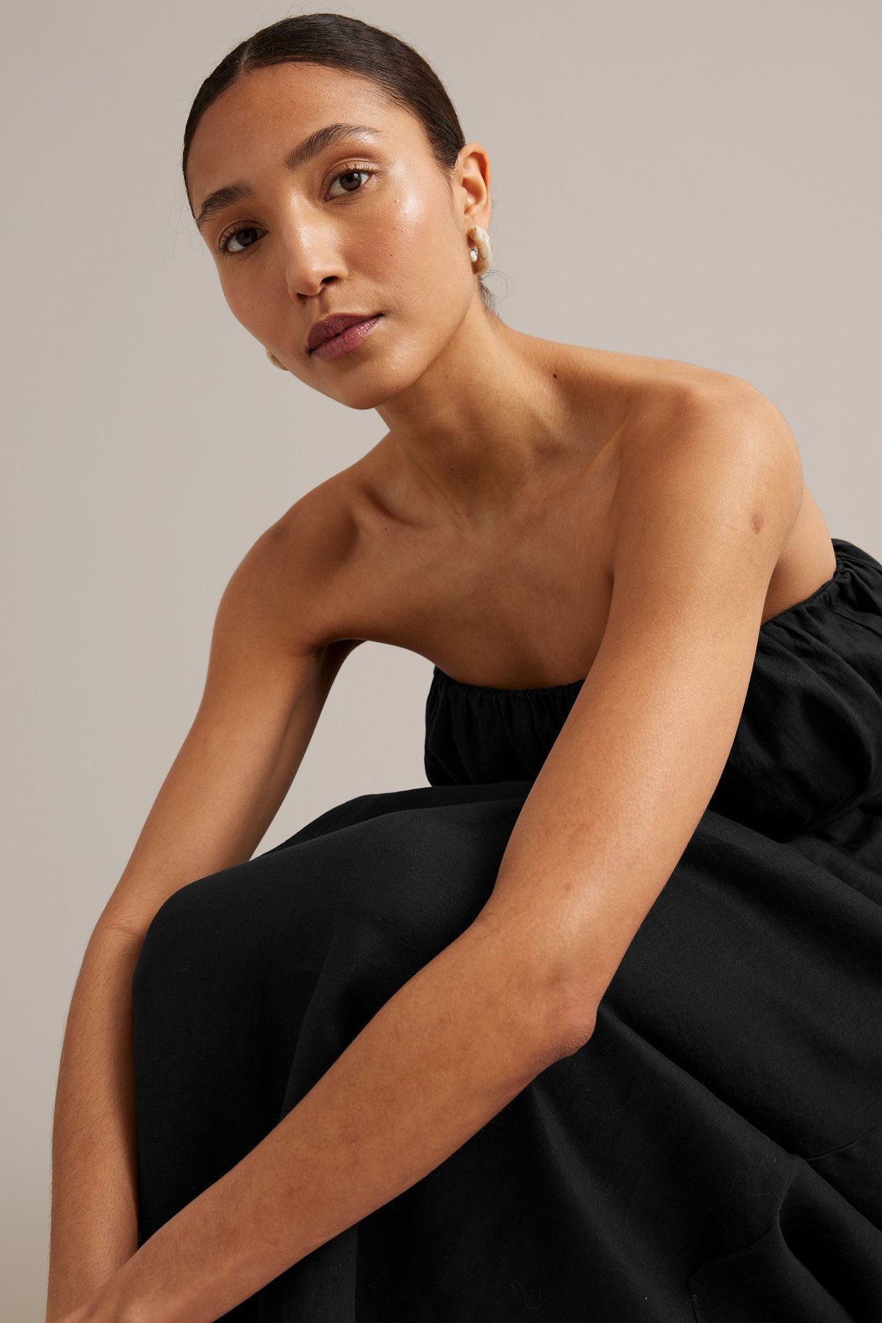 Närbild framifrån på en kvinna i en svart axelbandslös linnklänning med rynkad halslinning och ljusa örhängen.