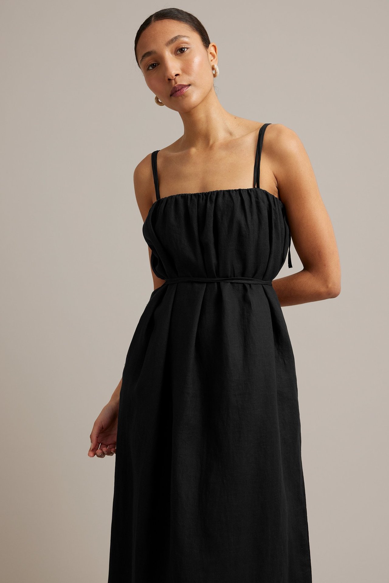 Forfra, kvinne i sort linkjole med spaghettistropper og knytebelte, stylet med ringøredobber.