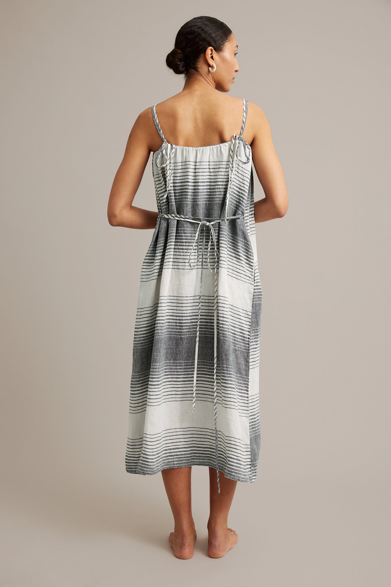 Kvinna bakifrån i randig linneklänning i mörkblått och vitt med tunna band och knutet midjebälte.