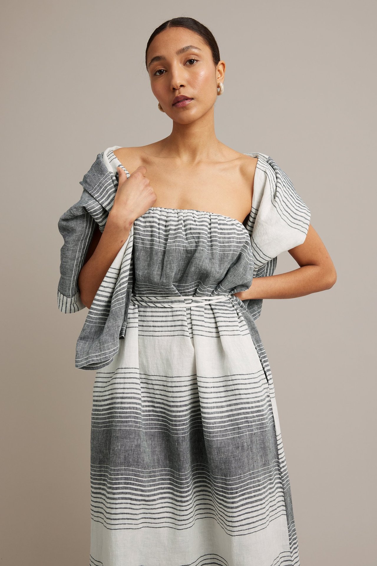 Framsidan av en kvinna i mörkgrå och vitrandig axelbandslös linneklänning med knytskärp.