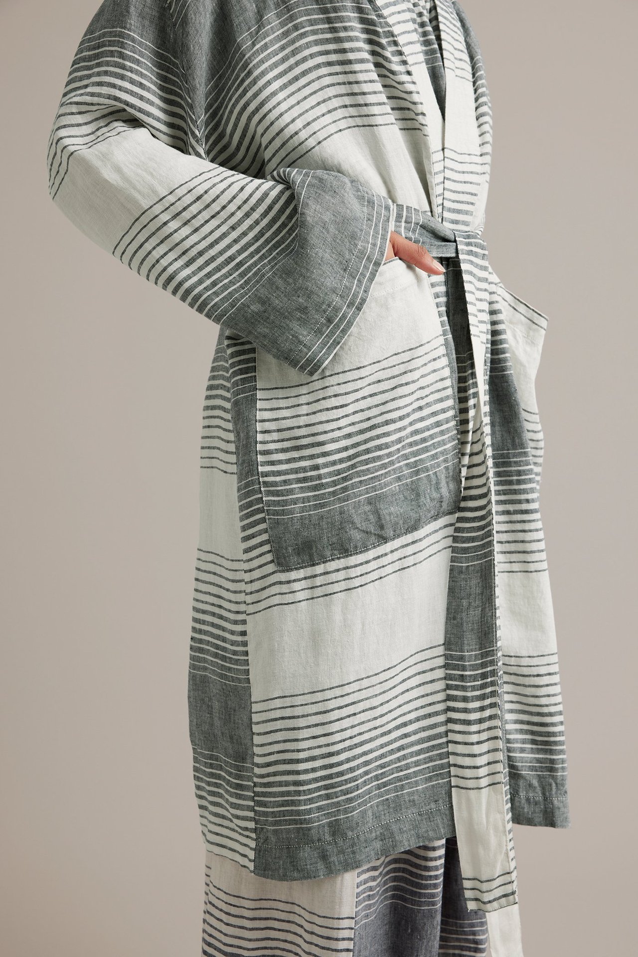 Sidovy: Kvinna i mörkgrå och naturvit randig linnekimono med knytskärp och ficka.