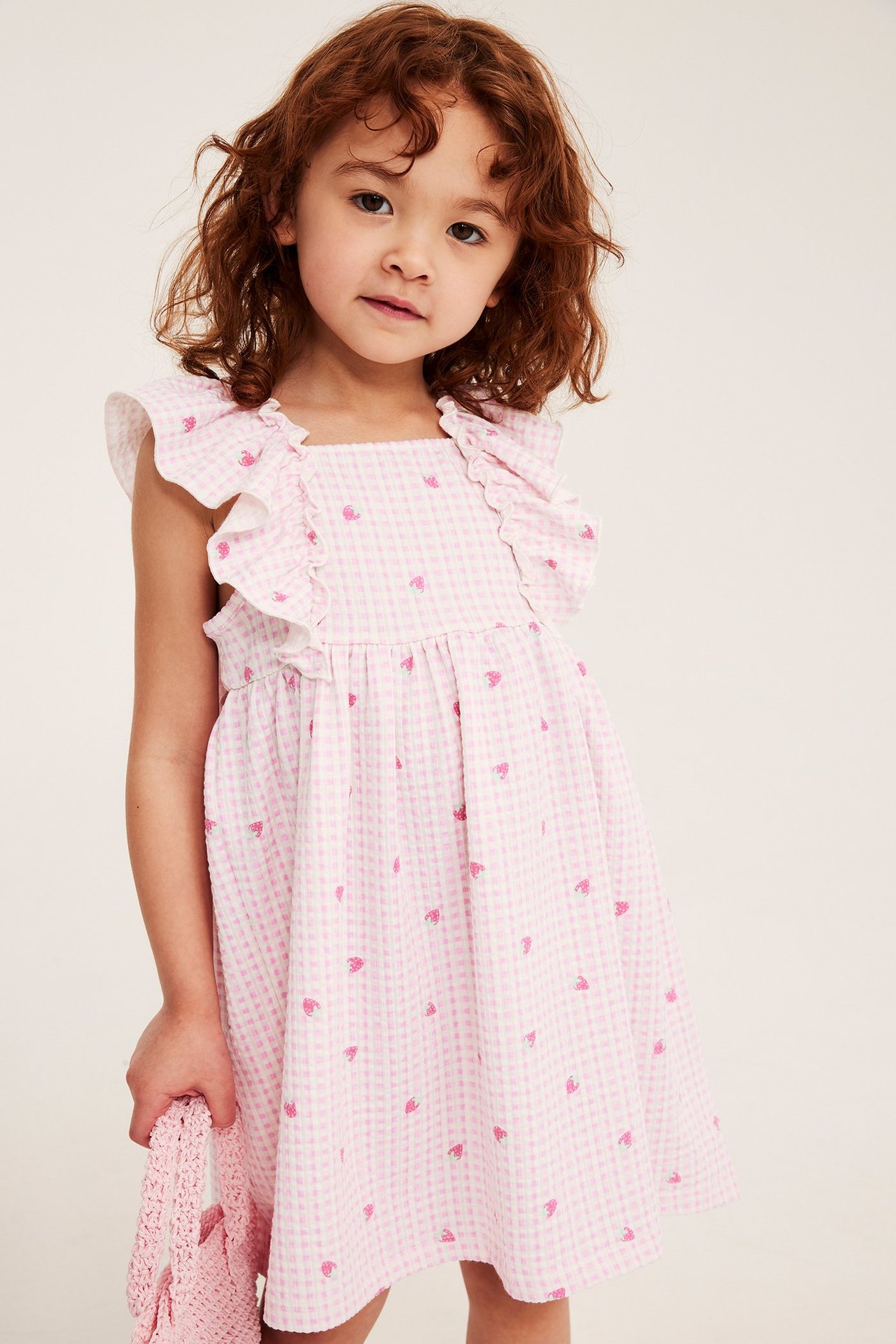 Edestä: lapsi vaaleanpunaisessa ruudullisessa mekossa mansikkakuviolla ja röyhelöillä, pitää virkattua laukkua.