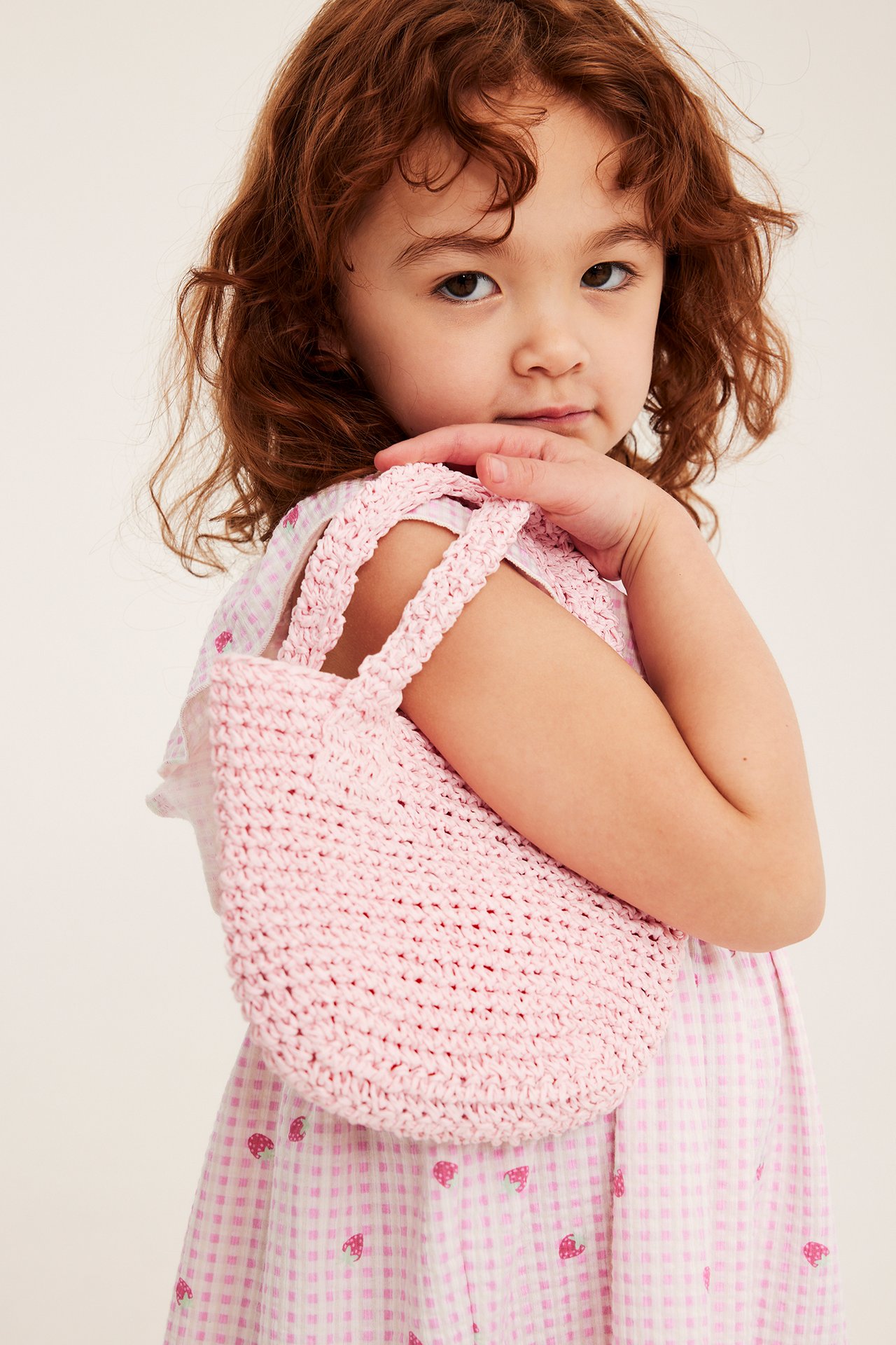 Zbliżenie na dziecko w jasnoróżowej sukience w kratę z truskawkowym wzorem, trzymające małą różową szydełkową torebkę.