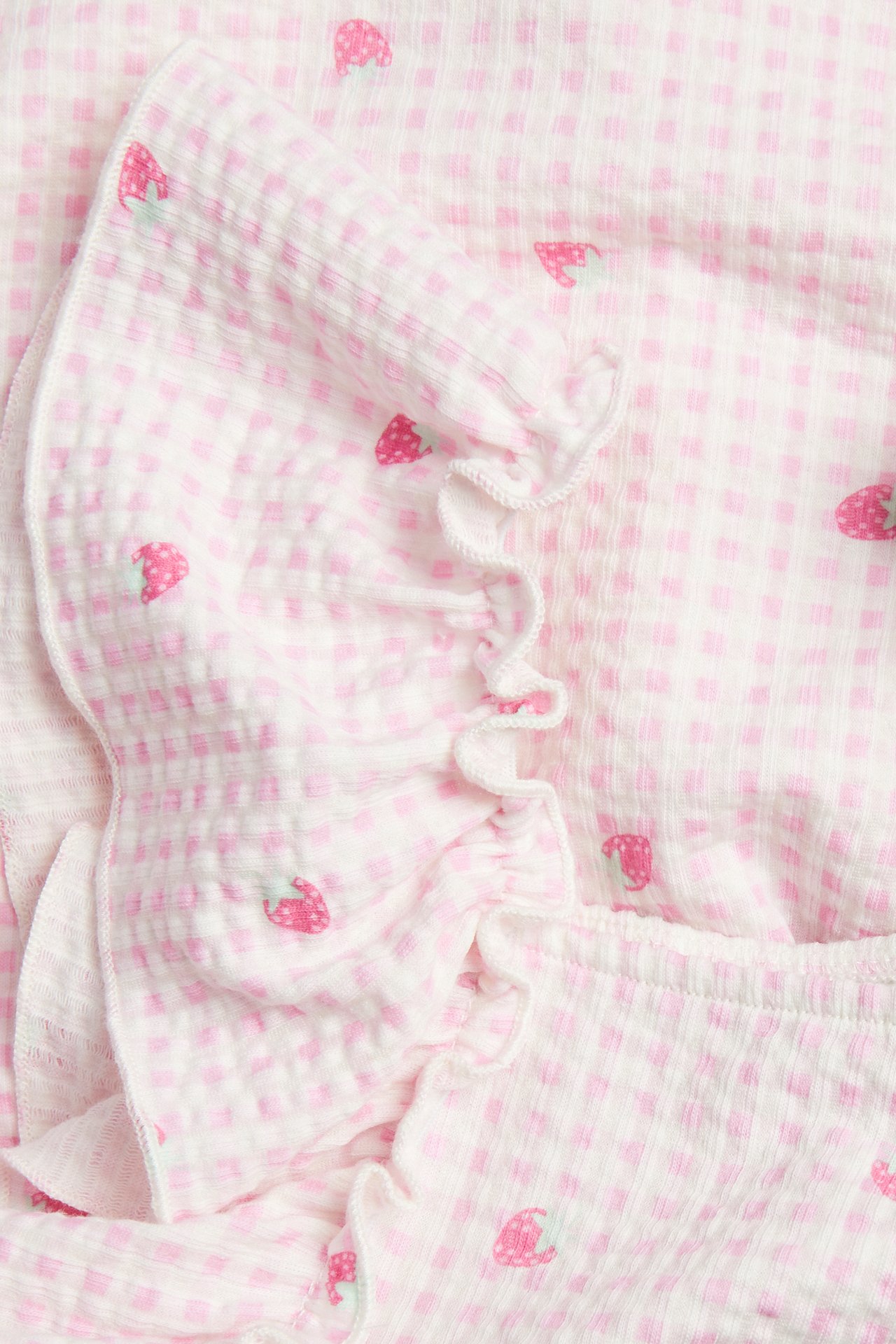 Zbliżenie na dziecięcą sukienkę z jasnoróżowej bawełny w kratkę z truskawkowym wzorem i falbanką.