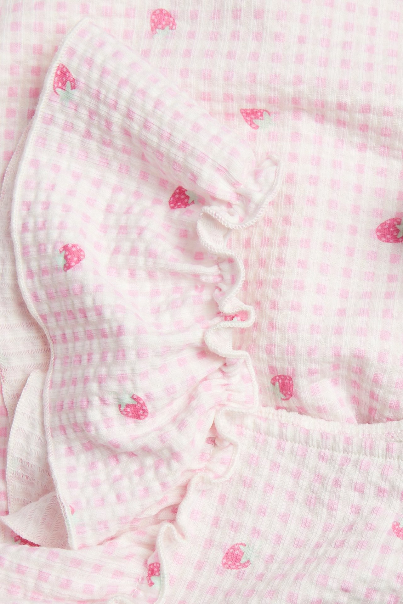 Zbliżenie na dziecięcą sukienkę z jasnoróżowej bawełny w kratkę z truskawkowym wzorem i falbanką.