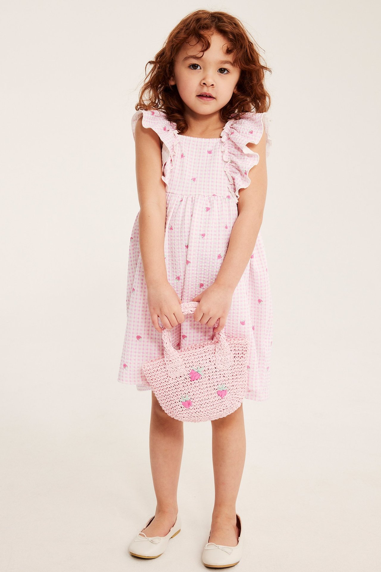 Edestäpäin: lapsi vaaleanpunaisessa ruudullisessa mansikkamekossa frillahioilla, pitelee pinkkiä laukkua.
