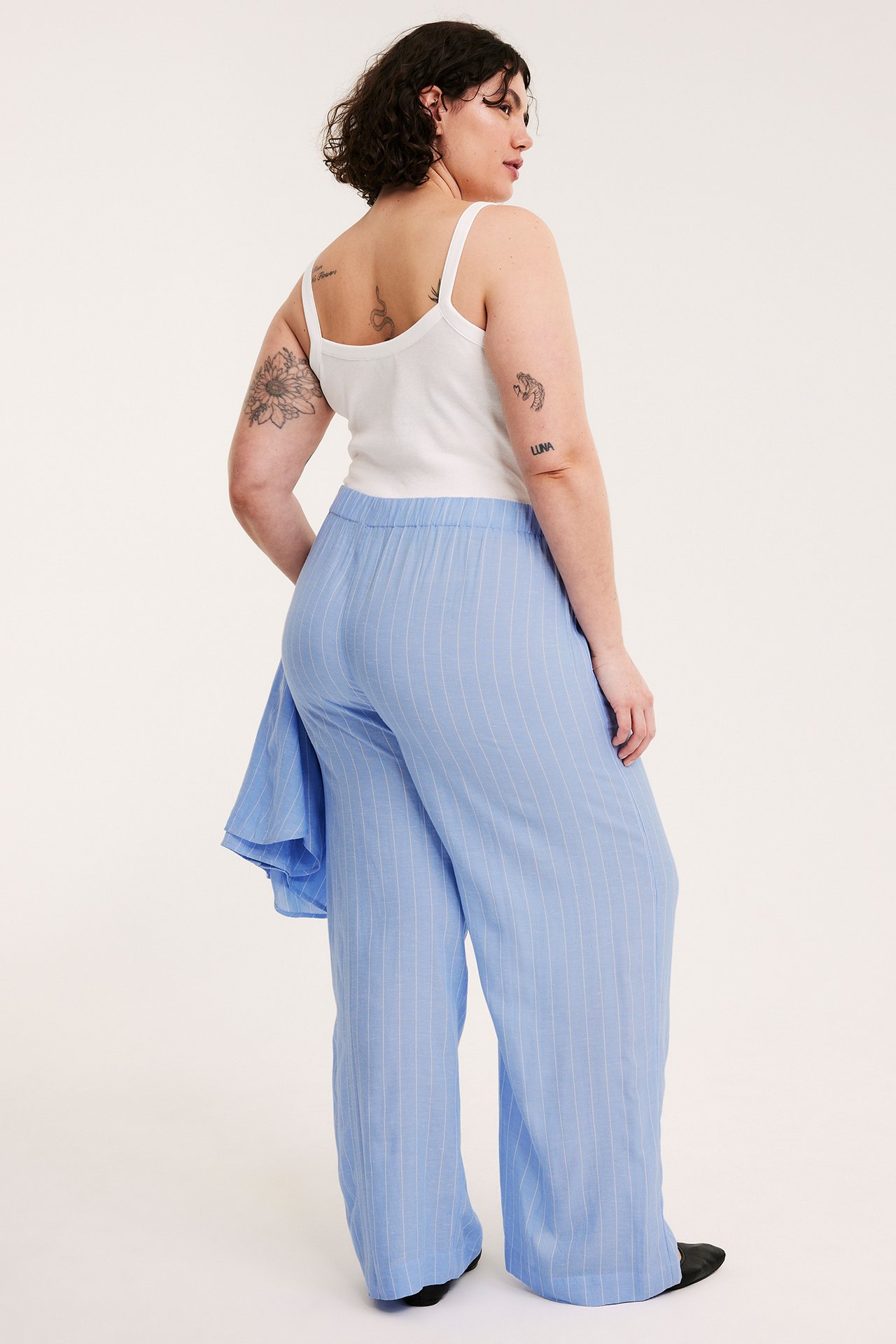 Nainen yllään vaaleansiniset raidalliset leveälahkeiset housut, valkoinen toppi ja sininen paita olalla.