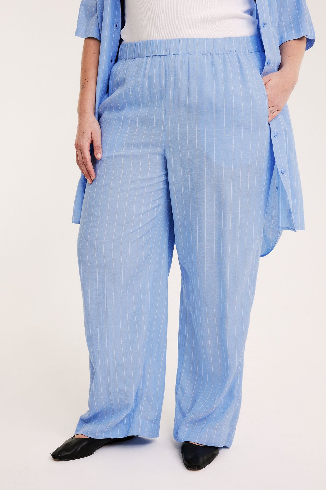 Edestä: Nainen vaaleansinisissä raidallisissa leveälahkeisissa housuissa, yhteensopivassa paidassa ja mustissa kengissä.