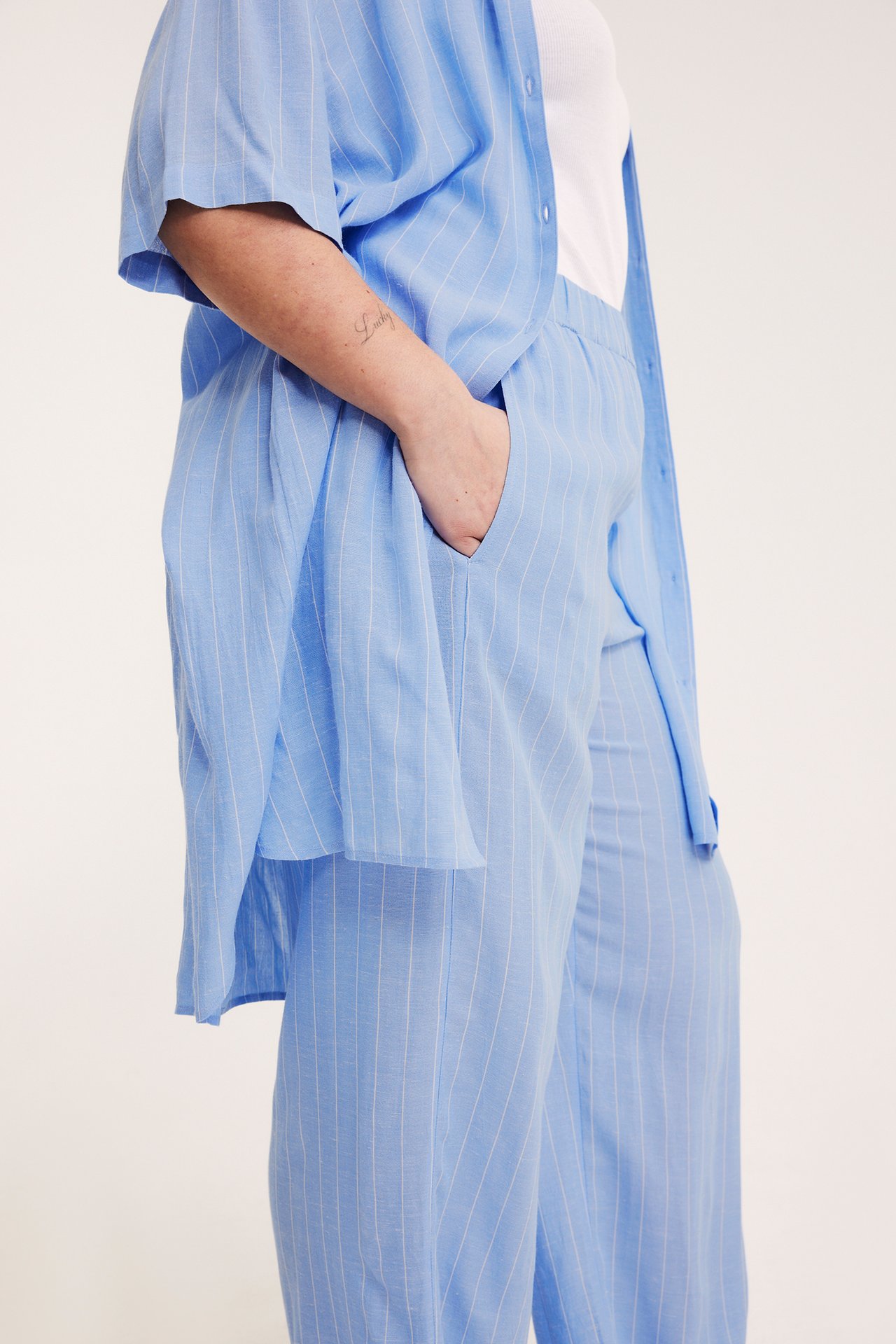 Nainen yllään vaaleansiniset raidalliset leveät housut, käsi taskussa, ja yhteensopiva avoin paita.