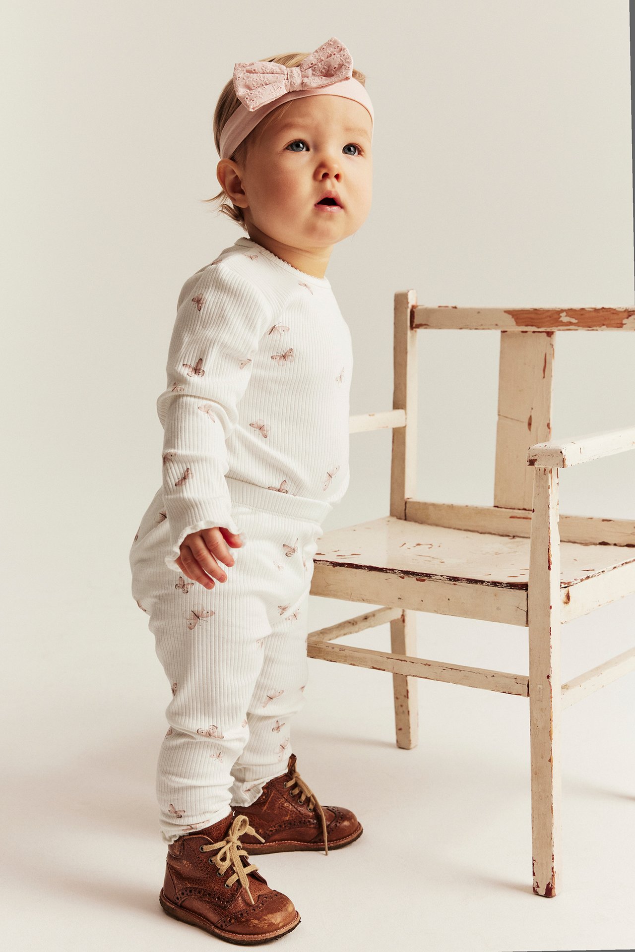 Sidevisning av en baby i offwhite ribbestrikkede sommerfuglmønstrede leggings, matchende topp, lyserosa pannebånd og brune sko.