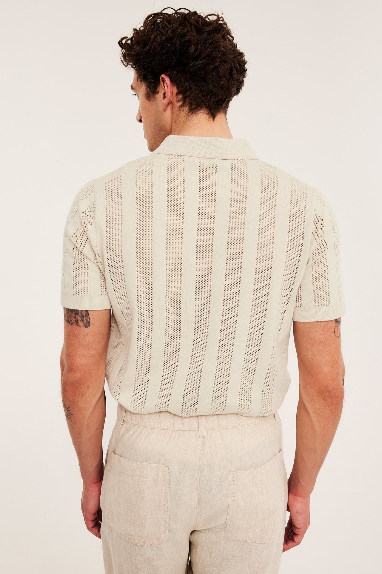 Mies takaa, yllään vaaleanbeige neulottu pikeepaita pystyraidallisella reikäkuviolla ja beiget housut.