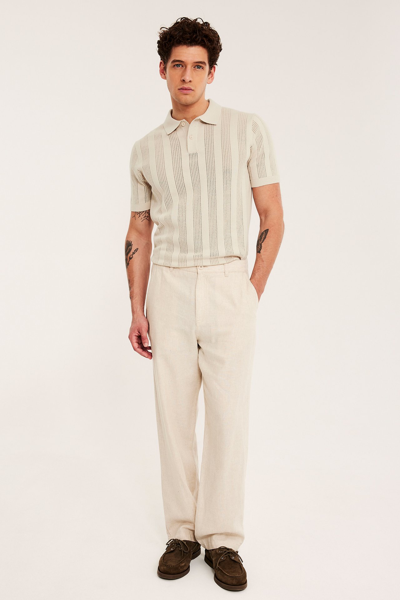 Framifrån: Man i ljusbeige stickad pikétröja, matchande byxor och mörkbruna båtskor.