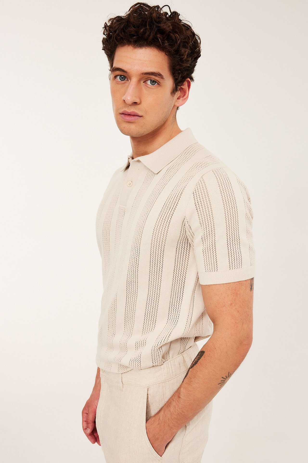 Mies yllään vaaleanbeige neulottu lyhythihainen pikeepaita, jossa kuviointi, ja vaaleanbeiget housut.