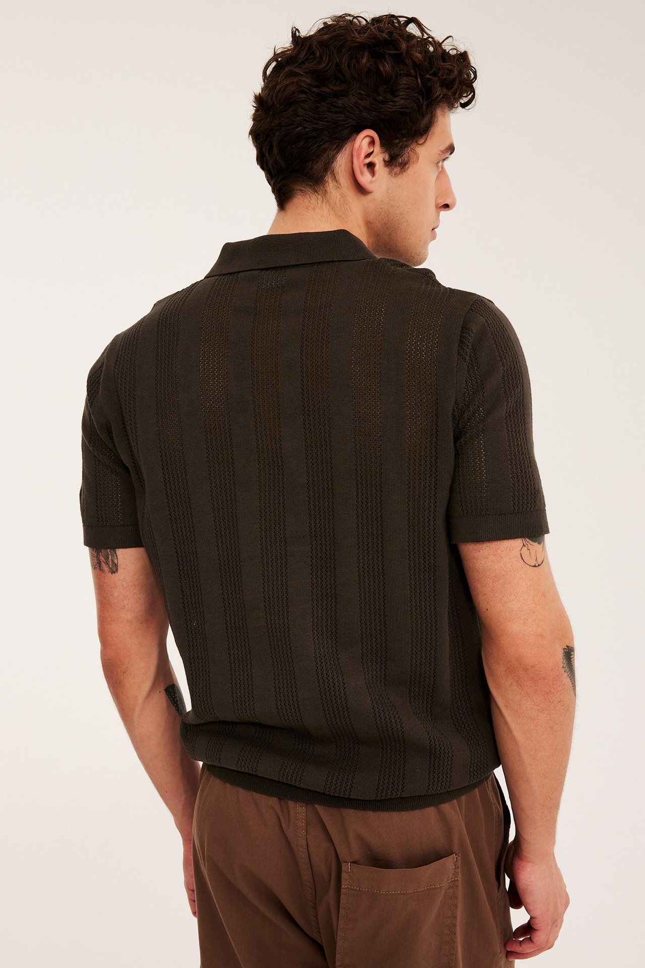 Mies takaa tummanruskeassa lyhythihaisessa pikeepaidassa pystysuoralla tekstuurilla ja ruskeissa housuissa.