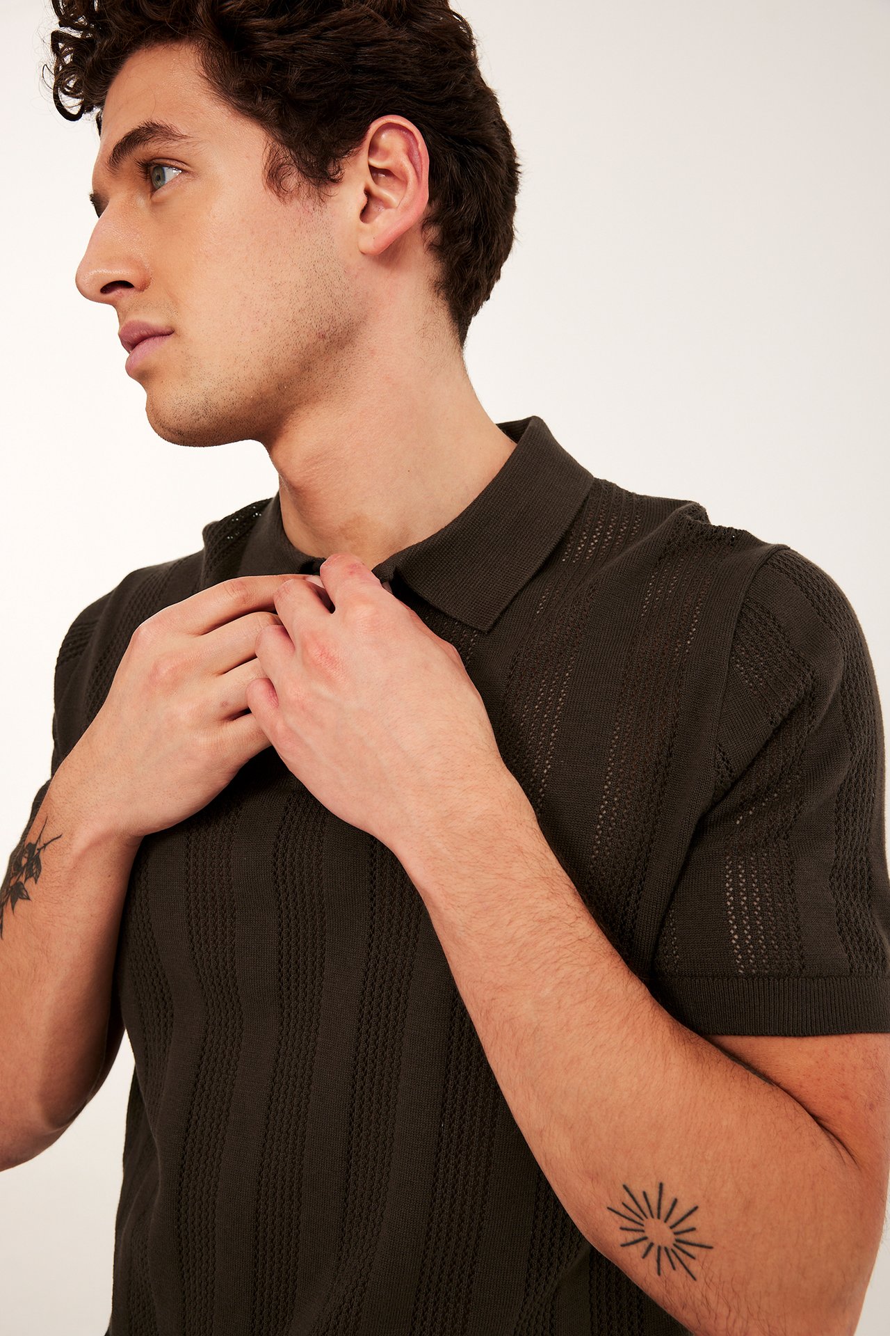 Närbild på man som justerar sin mörkbruna stickade pikétröja med vertikalt hålmönster och två knappar.