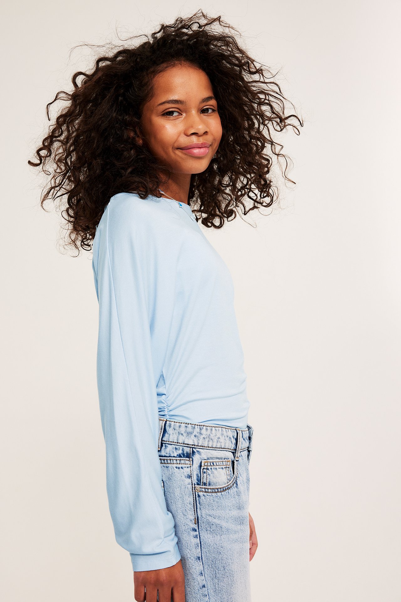 Sidovy av ett barn i en ljusblå långärmad topp med rynkad sida och ljusa jeans.